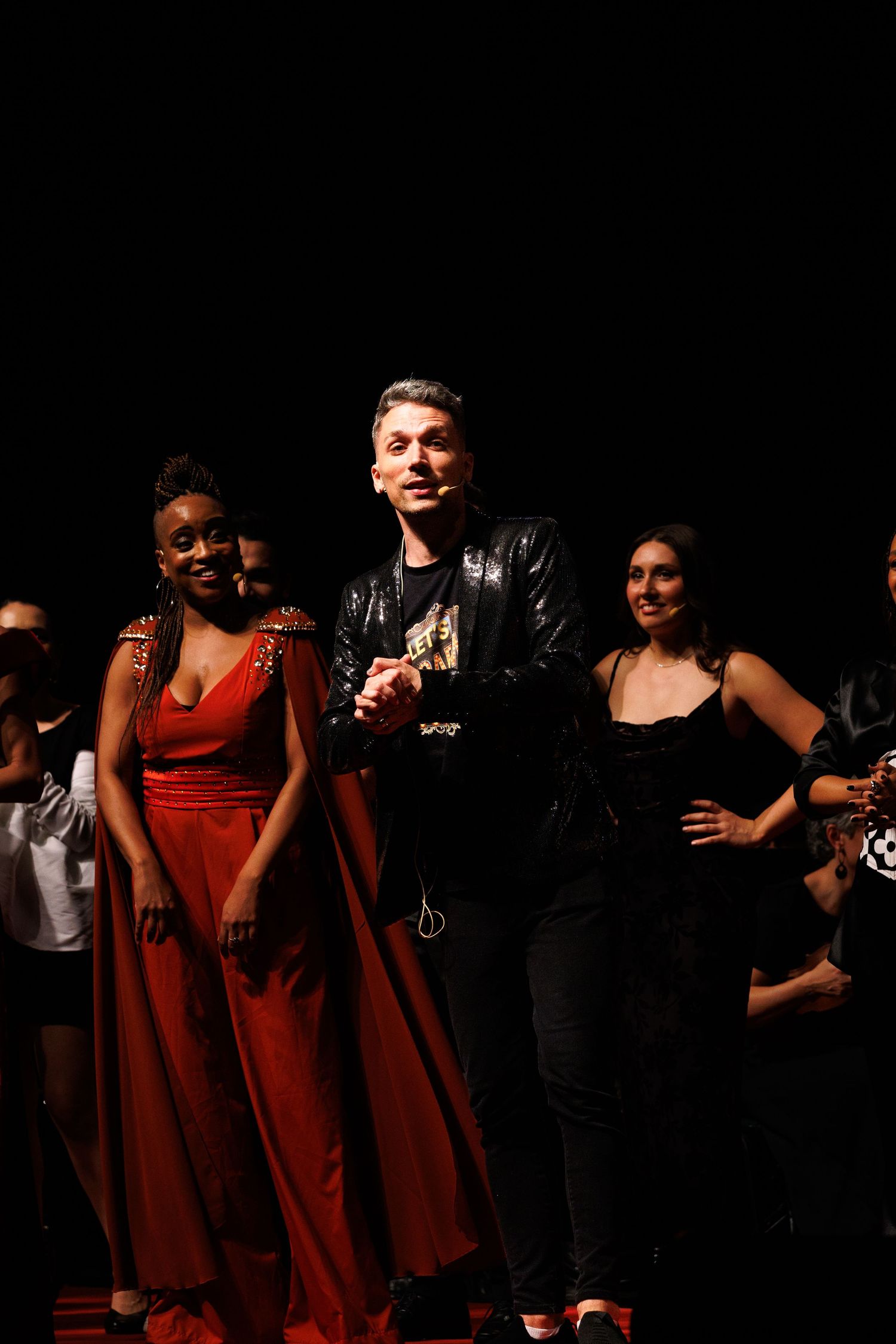 Intérprete de vestido vermelho ao lado de outro de jaqueta preta, com o elenco ao fundo.