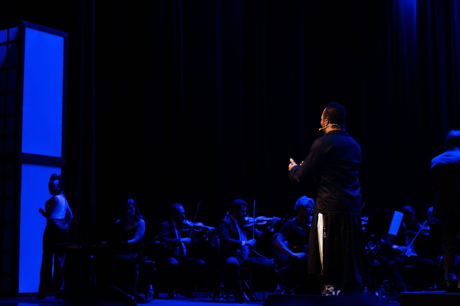 Plano aberto do palco escuro, com um solista ao centro e a orquestra sob luz azul.