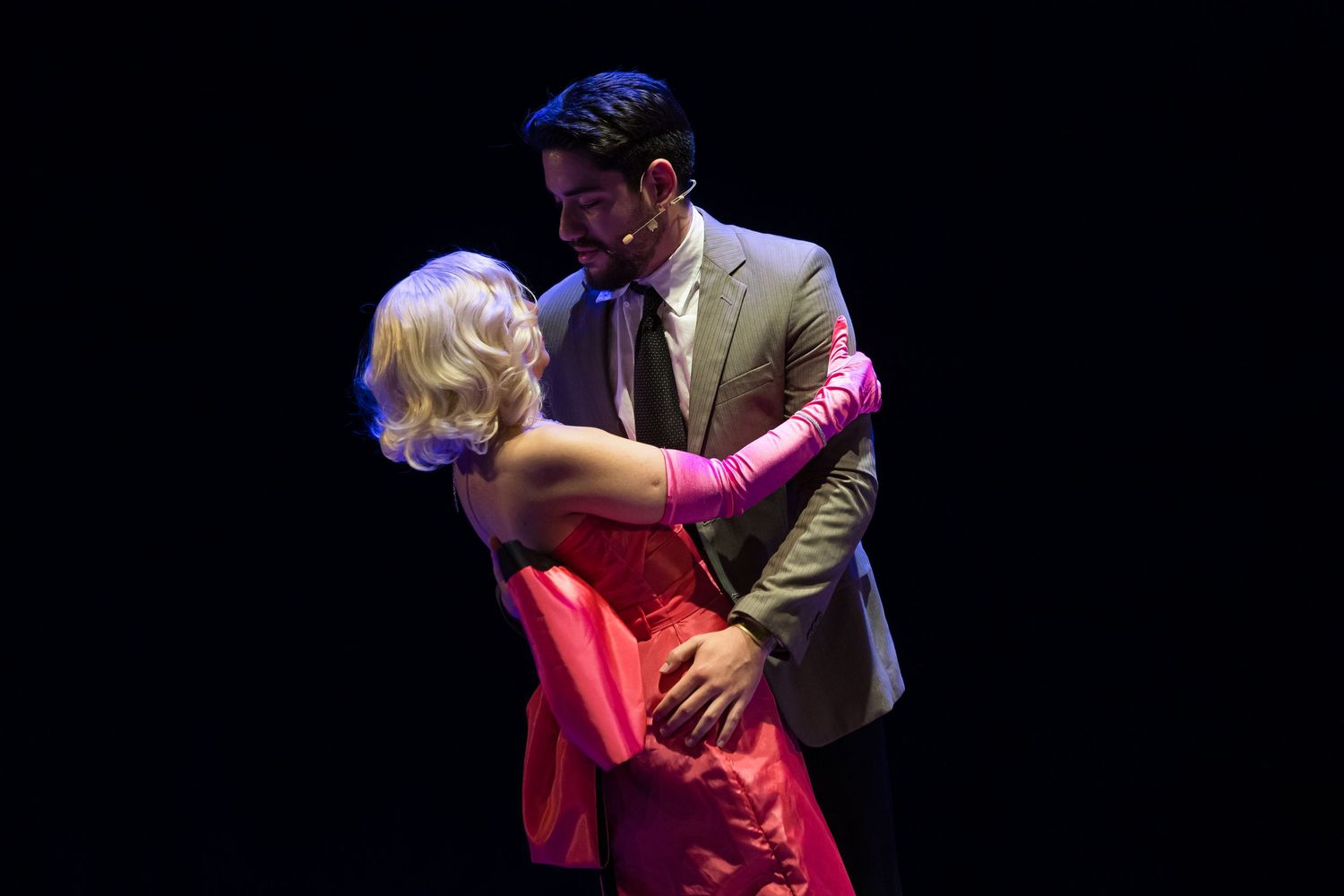 Casal em cena: intérprete de vestido rosa abraçada a outro de terno claro.