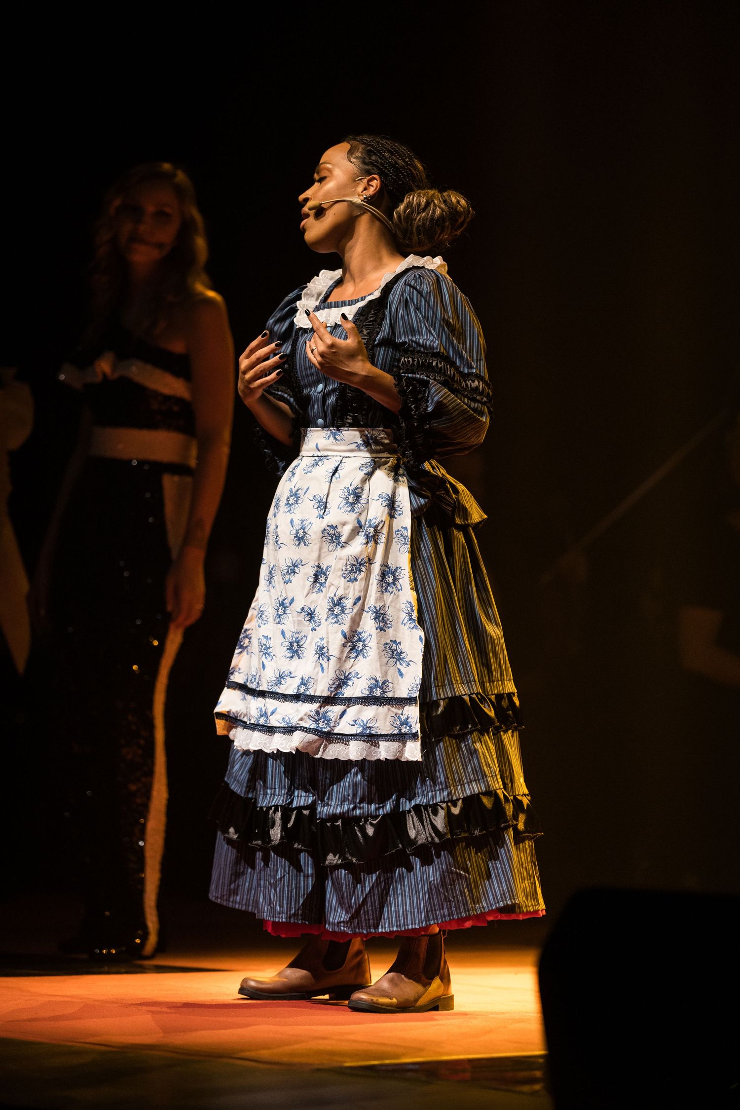 Intérprete de vestido de época azul e avental floral olha para cima, sob luz quente.