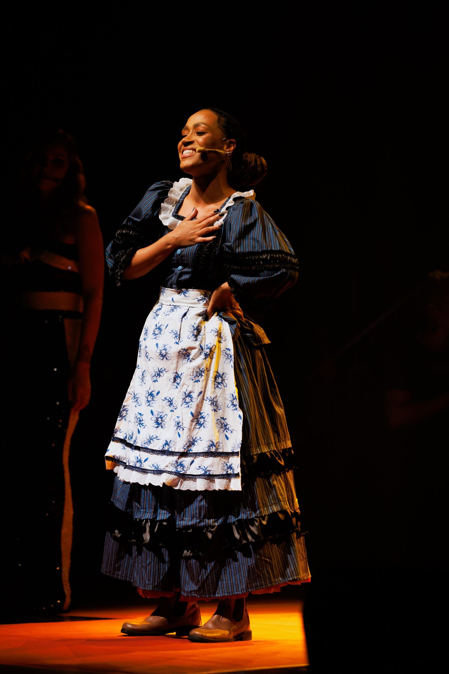 Intérprete de vestido de época azul e avental floral leva a mão ao peito, sobre o chão iluminado de laranja.