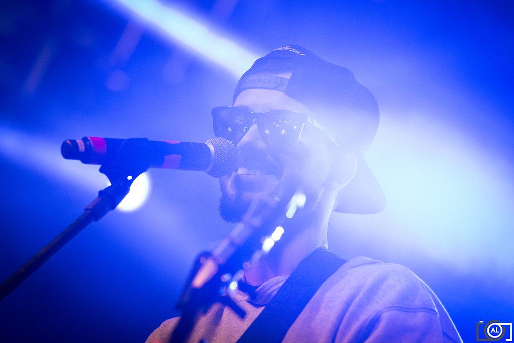 Cantor de boné e óculos escuros ao microfone sob luz azul.