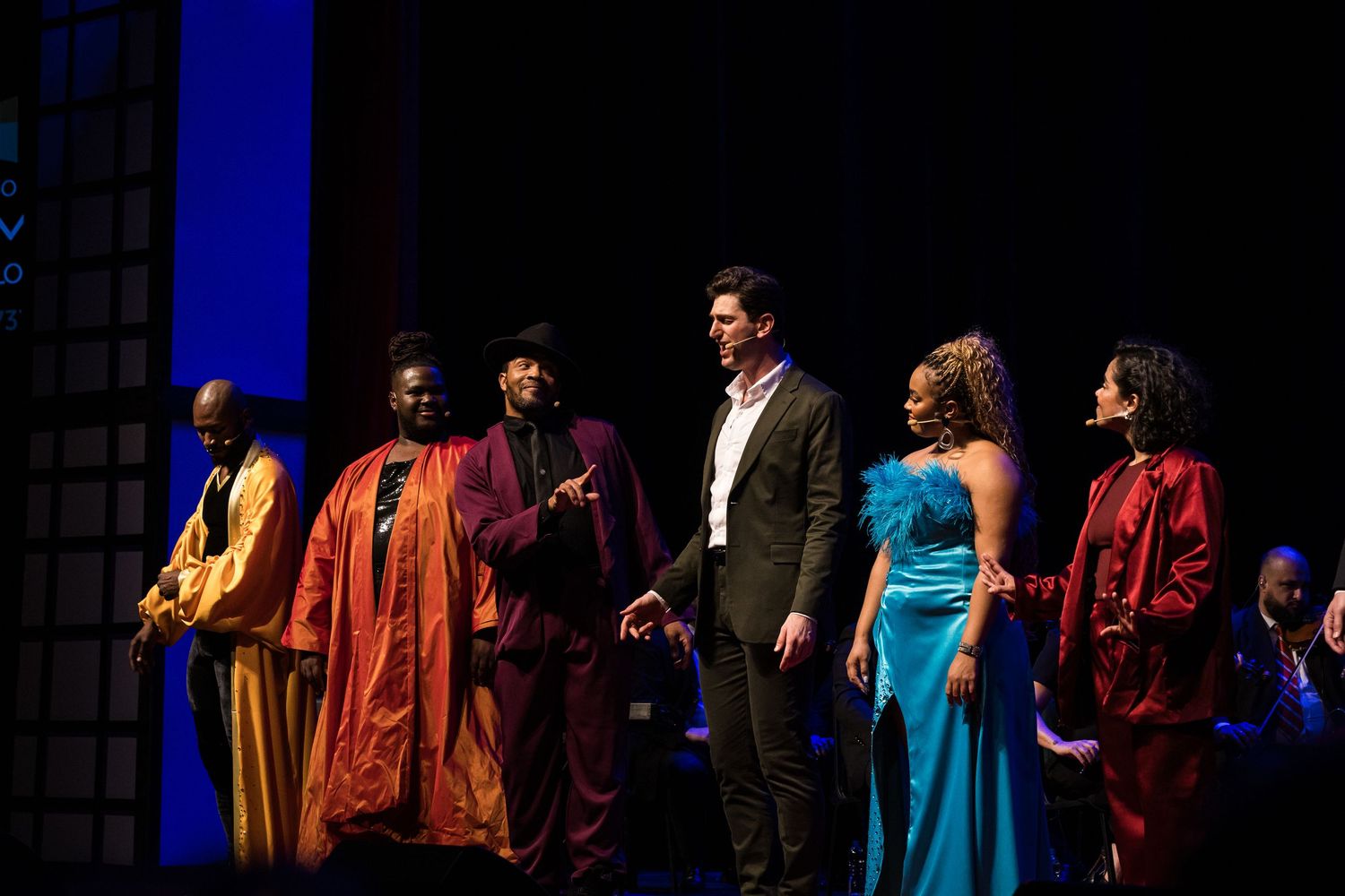 Elenco reunido no palco em figurinos coloridos diante de fundo azul.
