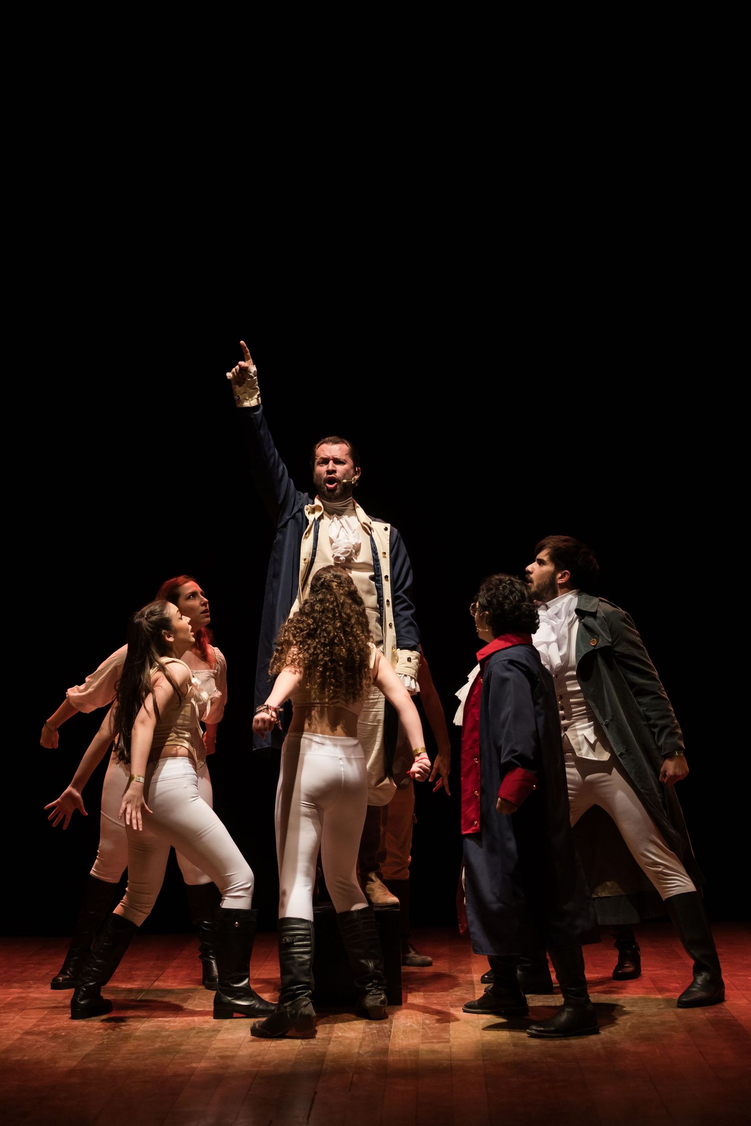 Ator em figurino branco ergue o braço no centro do palco, cercado pelo elenco, no musical Hamilton.