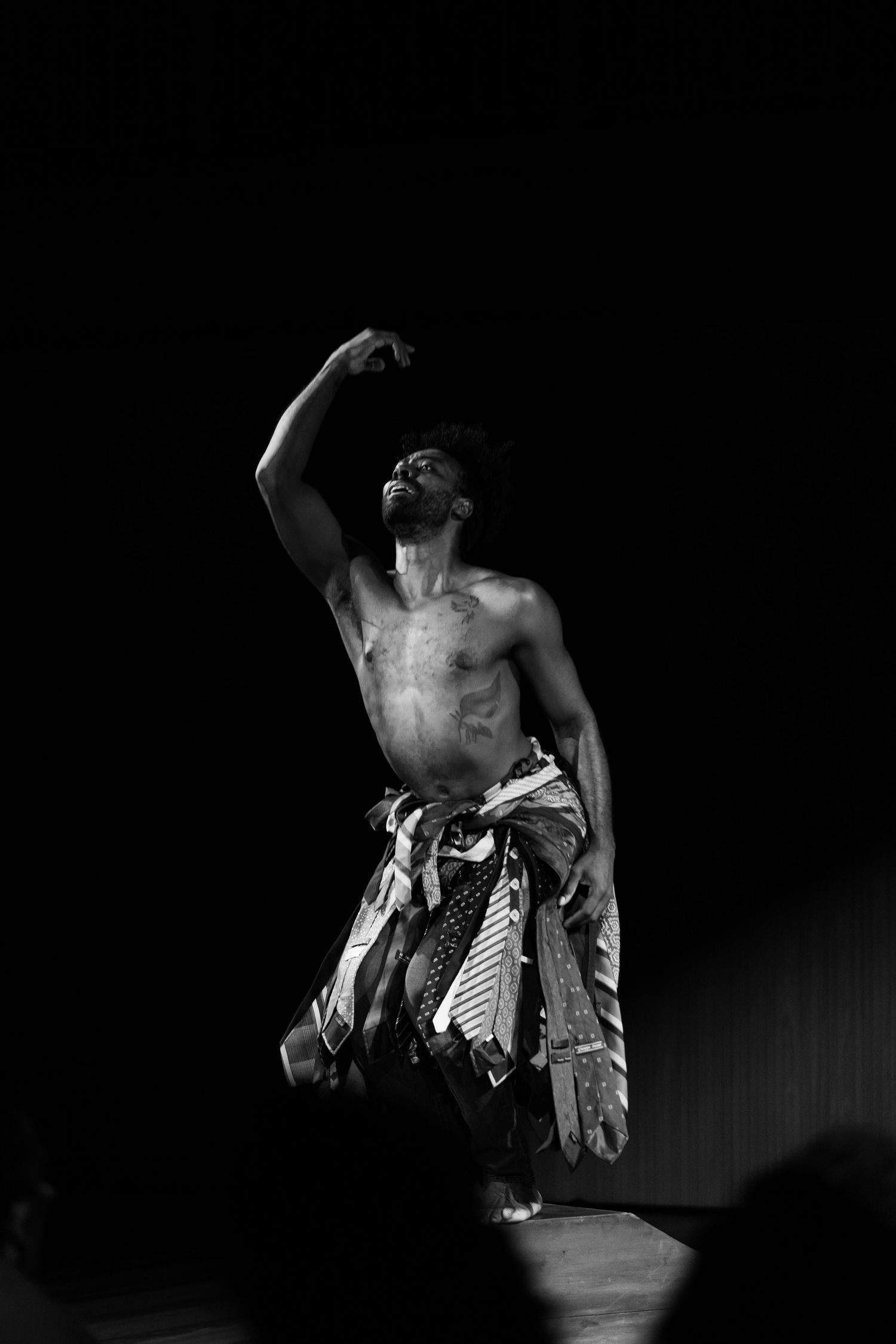 Intérprete sem camisa ergue o braço acima da cabeça, com saia de gravatas, em preto e branco.