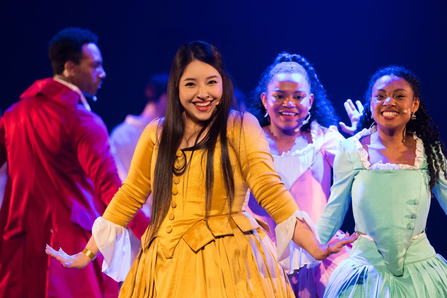 Três intérpretes em vestidos de época amarelo, azul-claro e verde sorriem, com o elenco ao fundo em luz azul.
