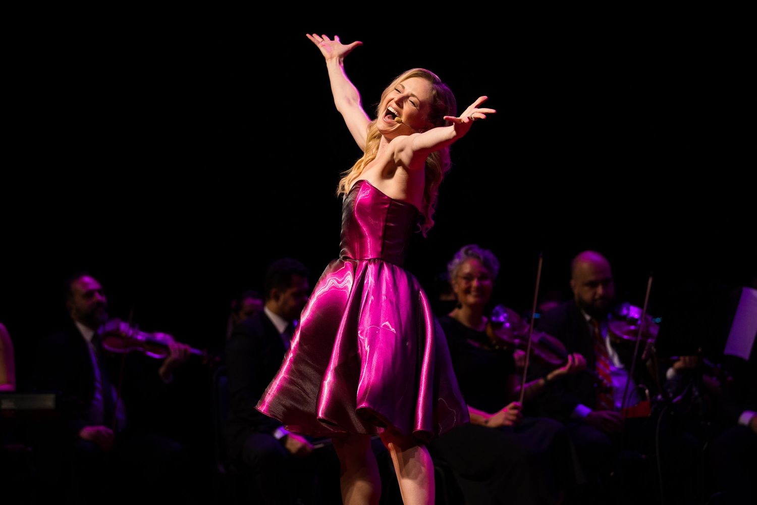 Intérprete de vestido rosa abre os braços diante da orquestra.