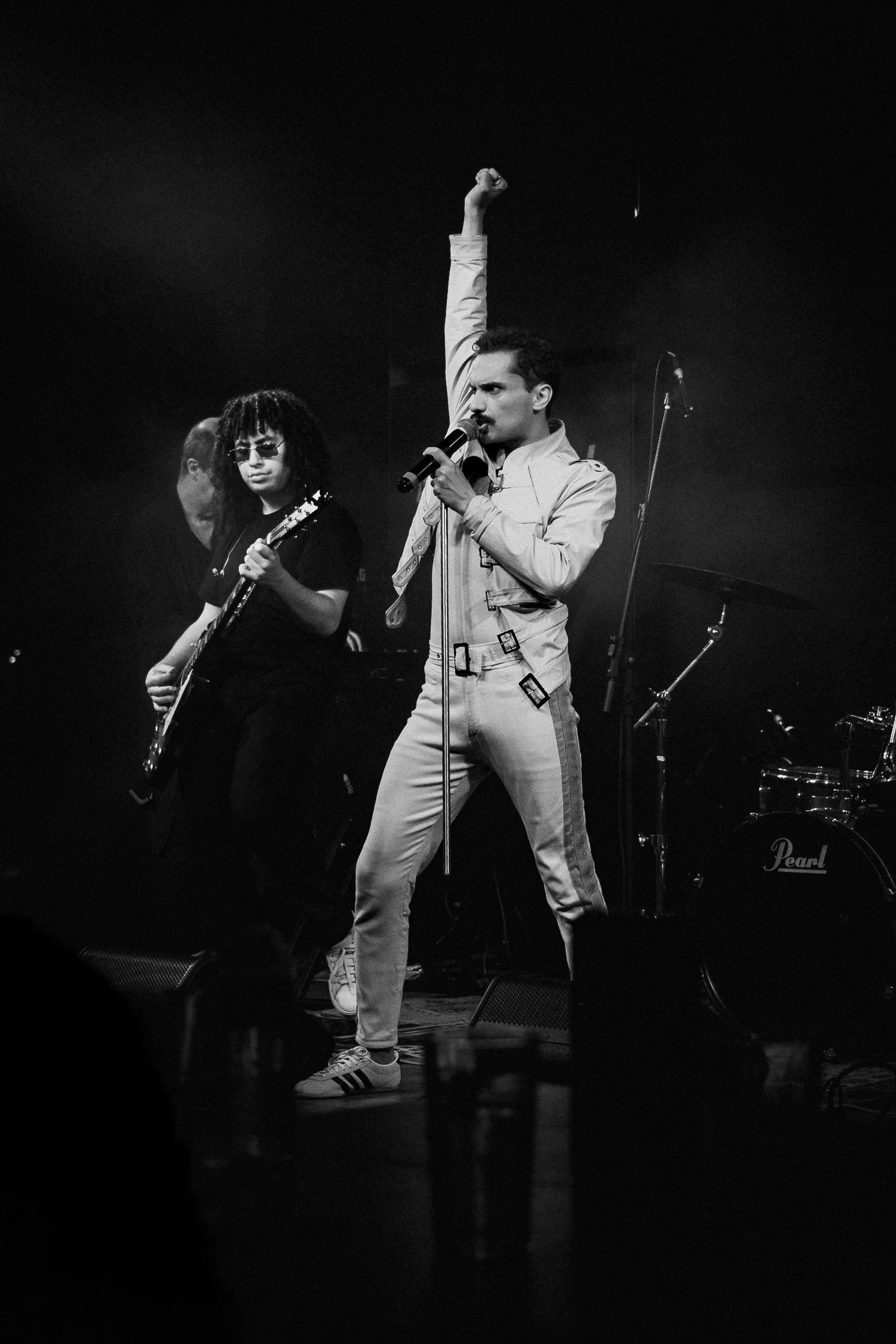 Vocalista de figurino branco ergue o punho ao microfone, com guitarrista atrás, em preto e branco.