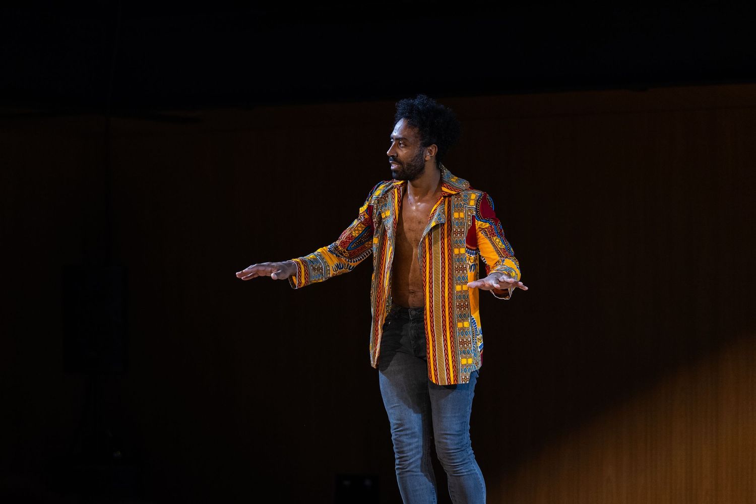 Intérprete de camisa estampada com os braços abertos, em plano aberto do palco de madeira.