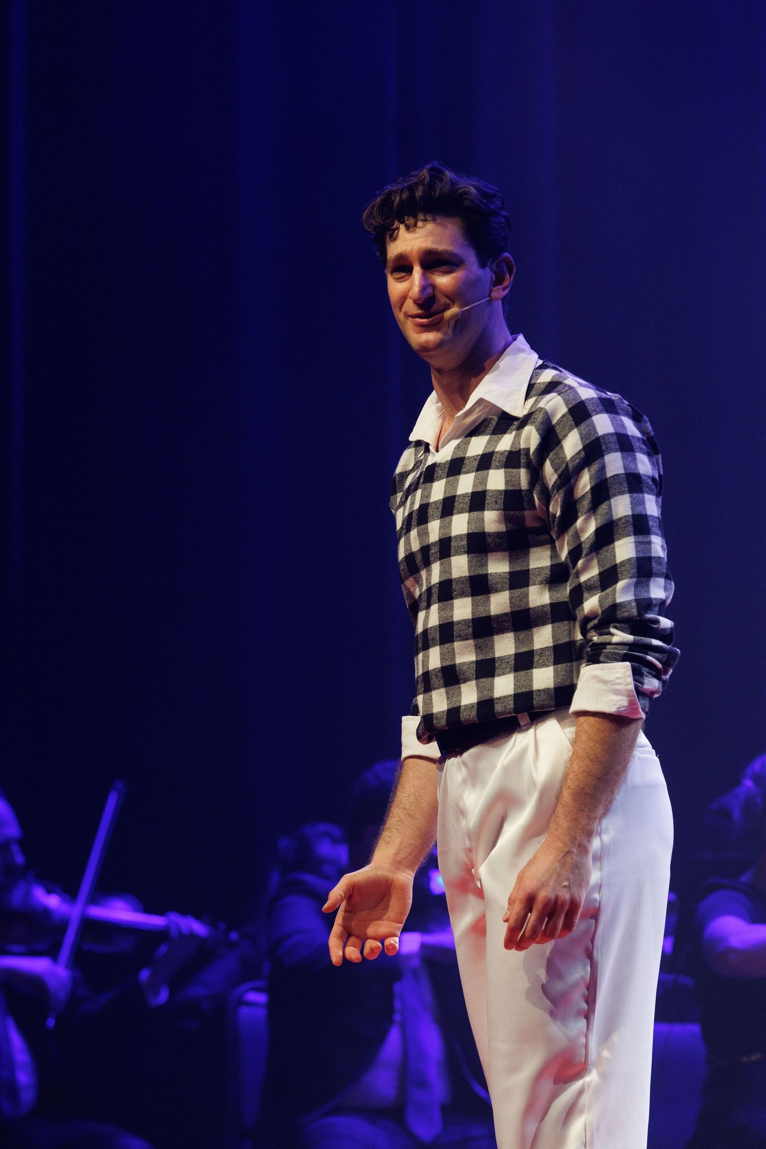 Intérprete de suéter xadrez preto e branco e calça branca em cena, com a orquestra ao fundo.