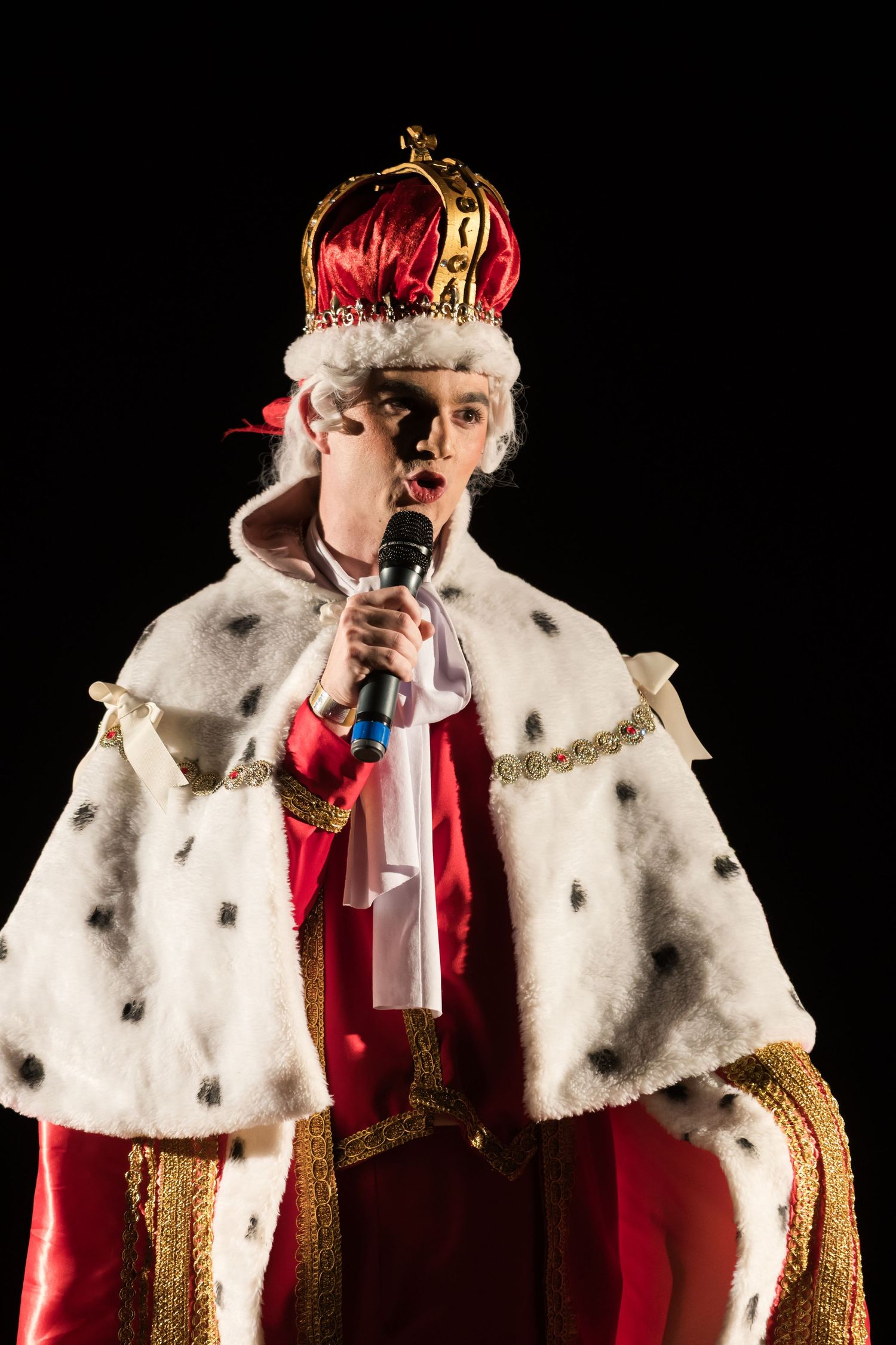 Intérprete do Rei George III, de coroa e manto de arminho, canta ao microfone.