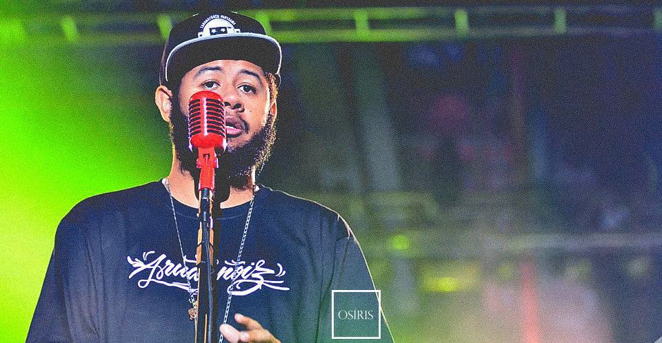 Rapper canta ao microfone sob luzes verdes e roxas.