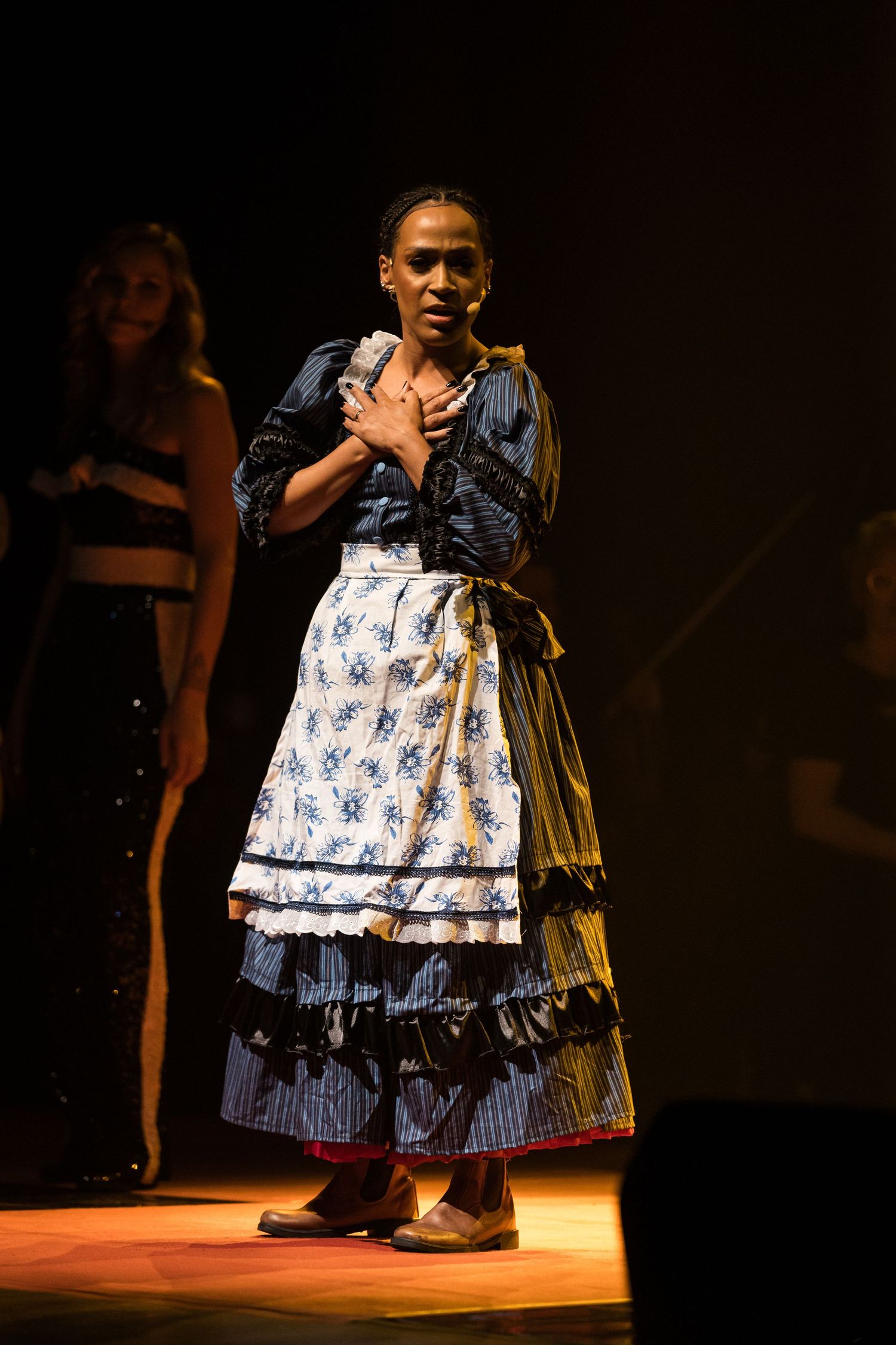 Intérprete de vestido de época azul e avental floral com as mãos no peito, sobre o chão iluminado de laranja.