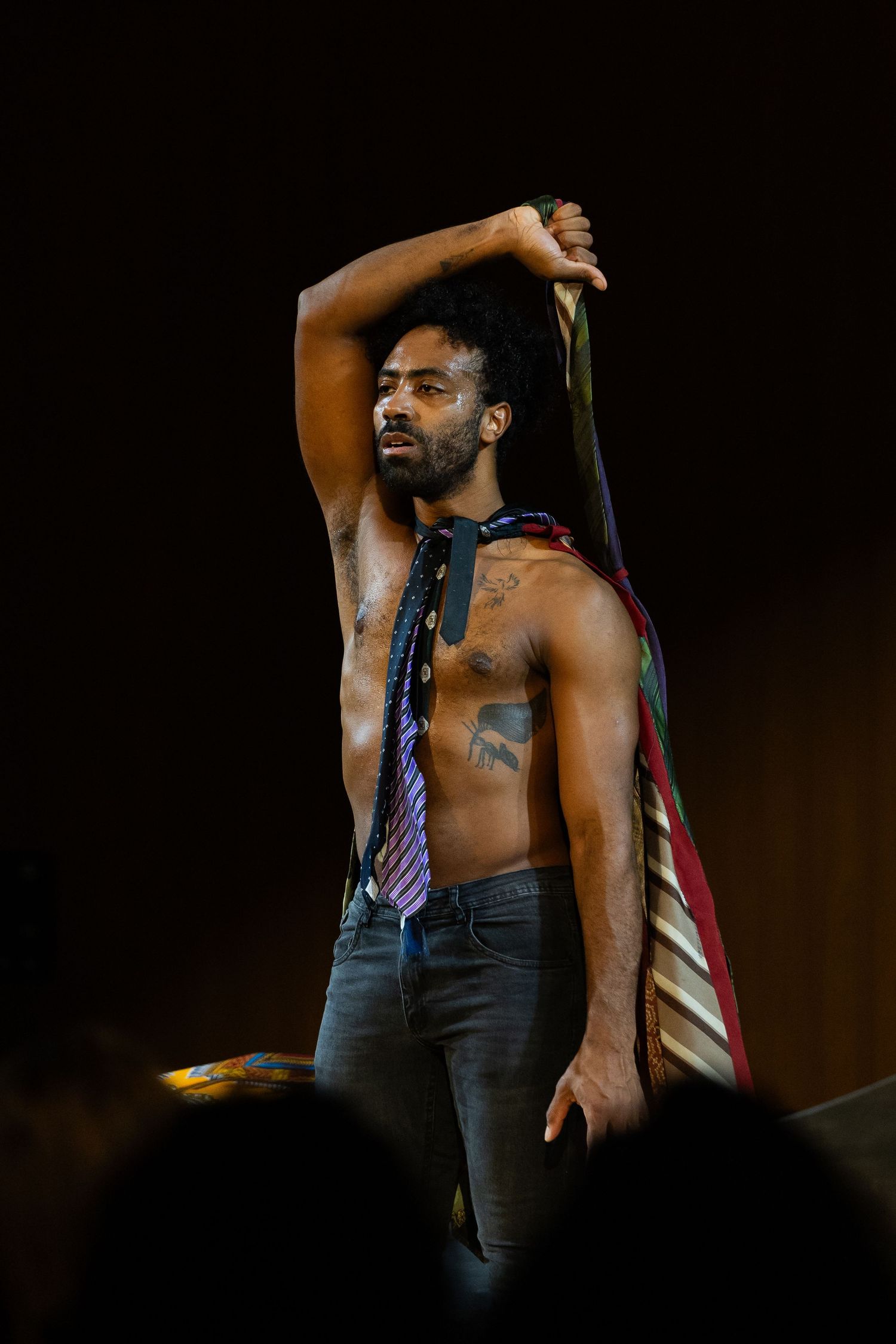 Intérprete sem camisa ergue o braço com um feixe de gravatas sobre a cabeça.