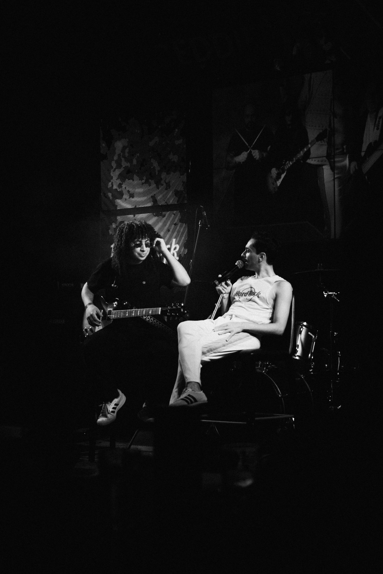 Guitarrista toca ao lado do vocalista sentado em um banco no palco, em preto e branco.