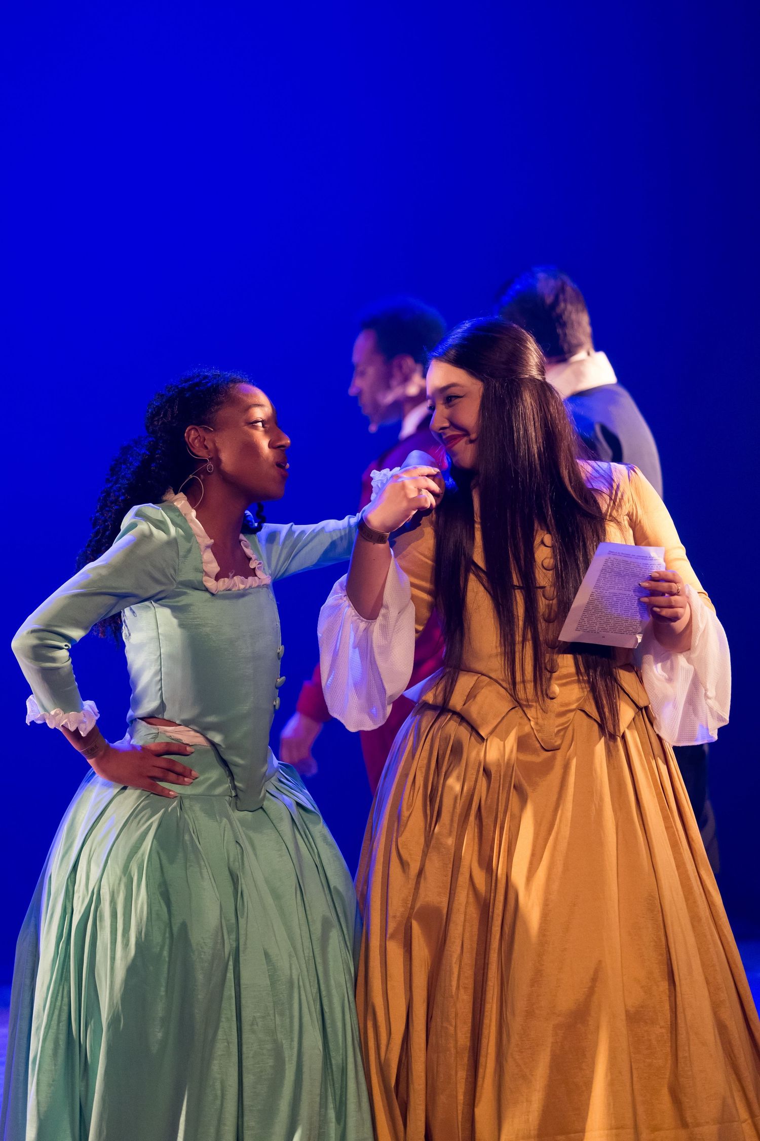 Duas intérpretes em vestidos de época verde e dourado conversam em cena, sob luz azul.