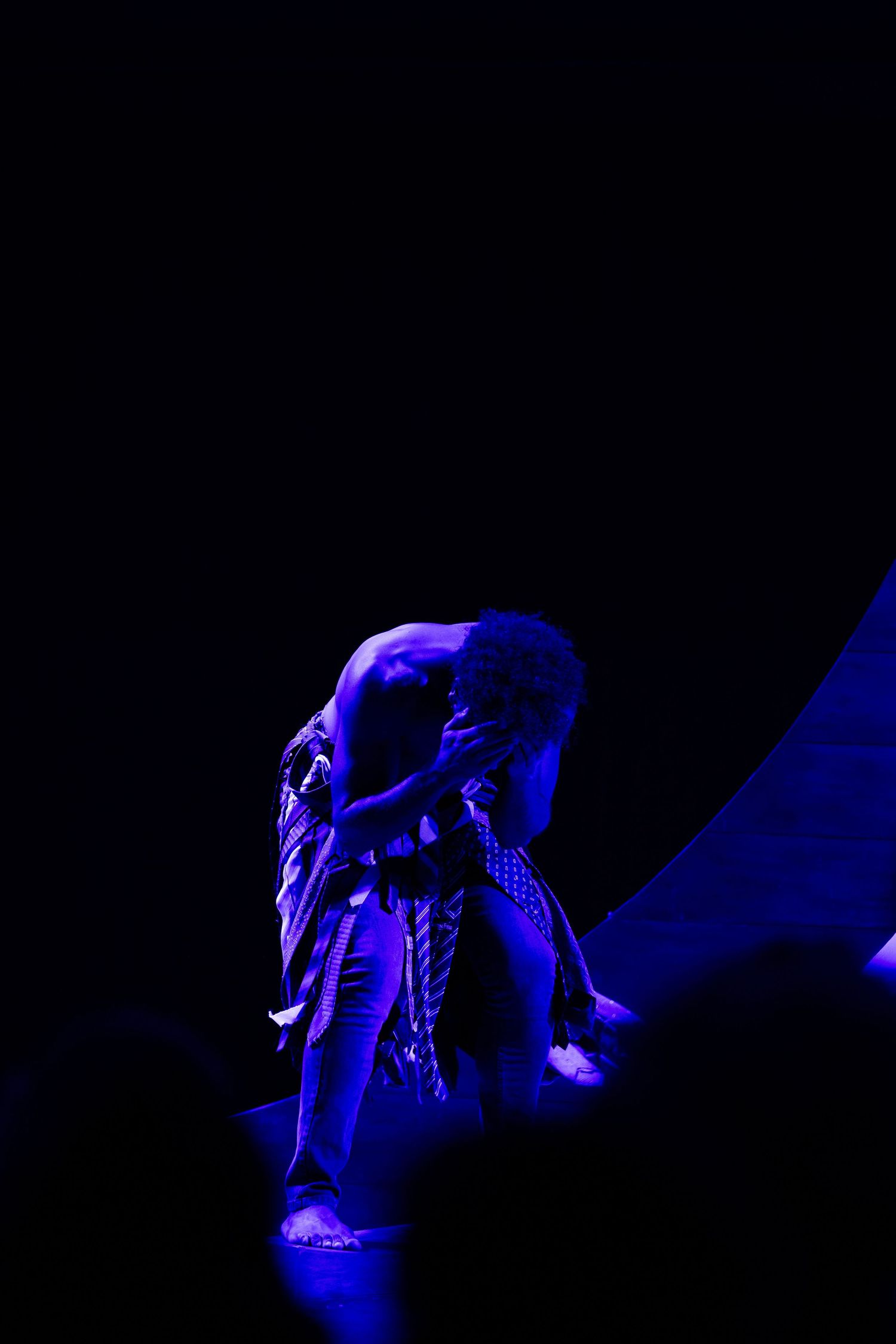 Vulto coberto de tecidos e gravatas, curvado sob luz azul.