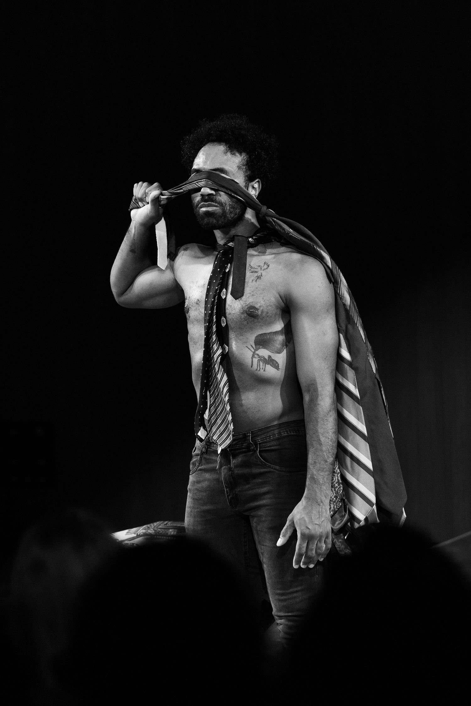 Ator sem camisa cobre o rosto com uma gravata, em fotografia de palco em preto e branco.