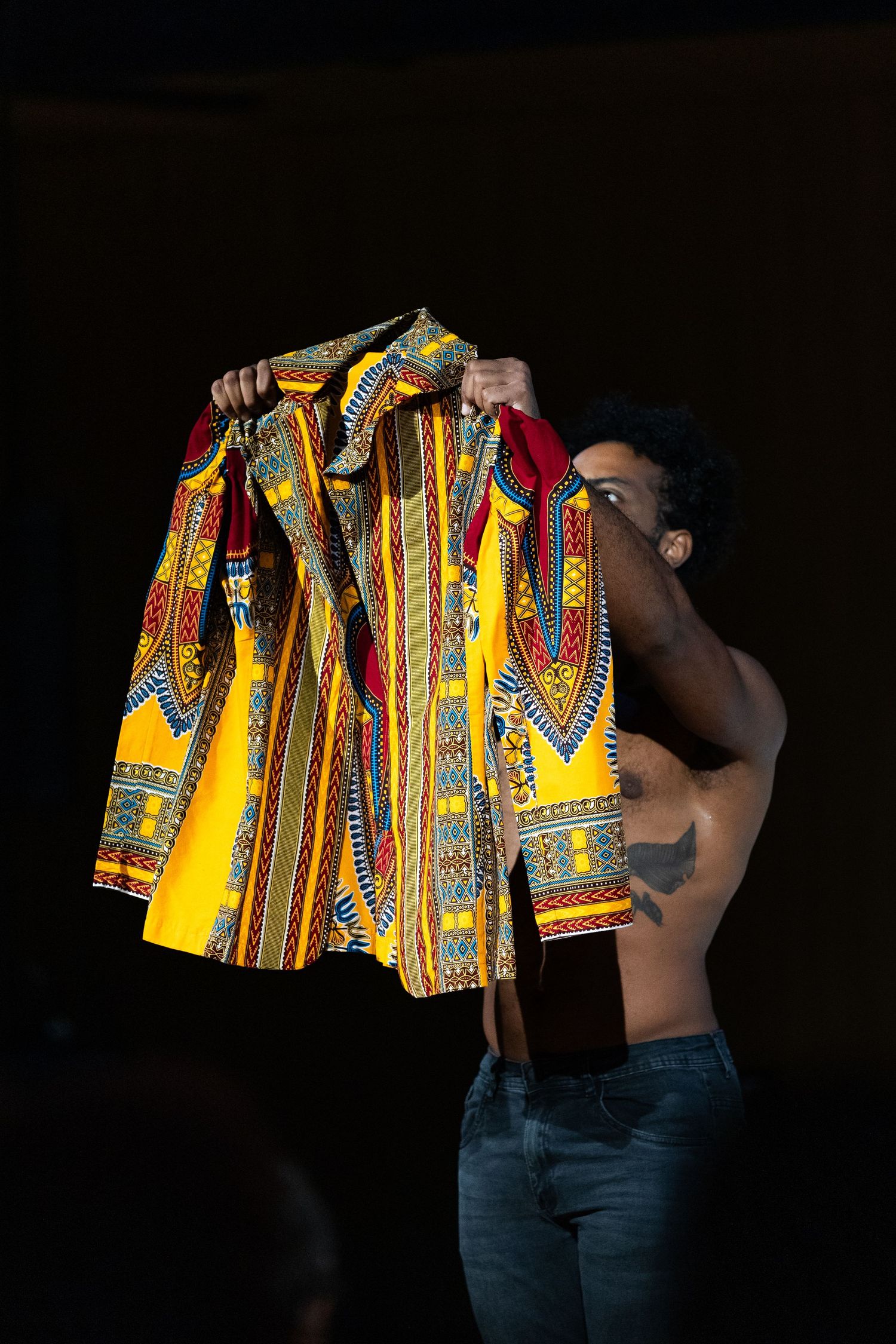 Intérprete sem camisa ergue uma camisa amarela de estampa africana diante do corpo.
