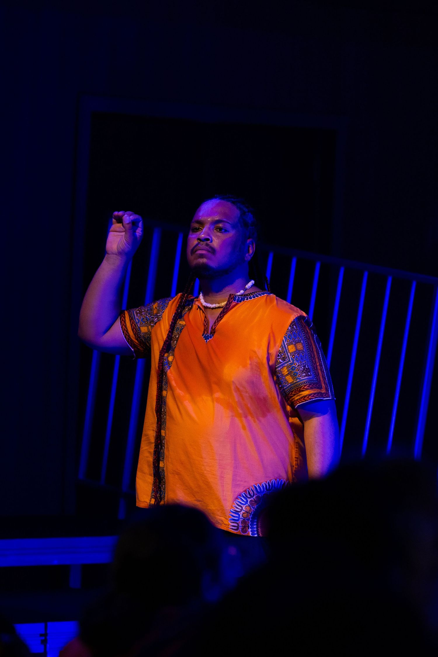 Intérprete de camisa laranja estampada ergue o punho, sob luz azul.