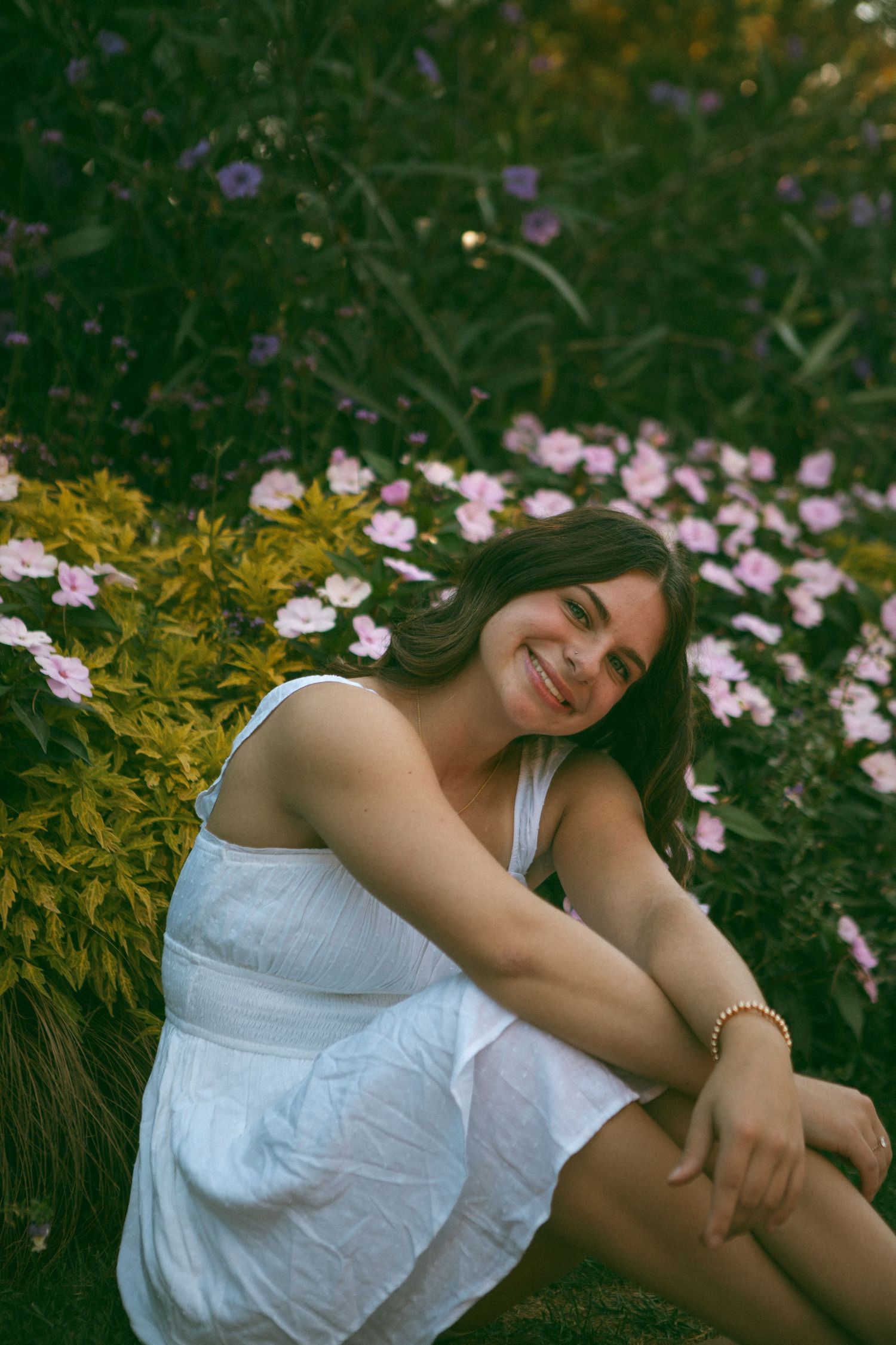 Smiling young woman in a white dress sitting among vibrant pink and purple flowers, radiating happiness in a serene garden setting.