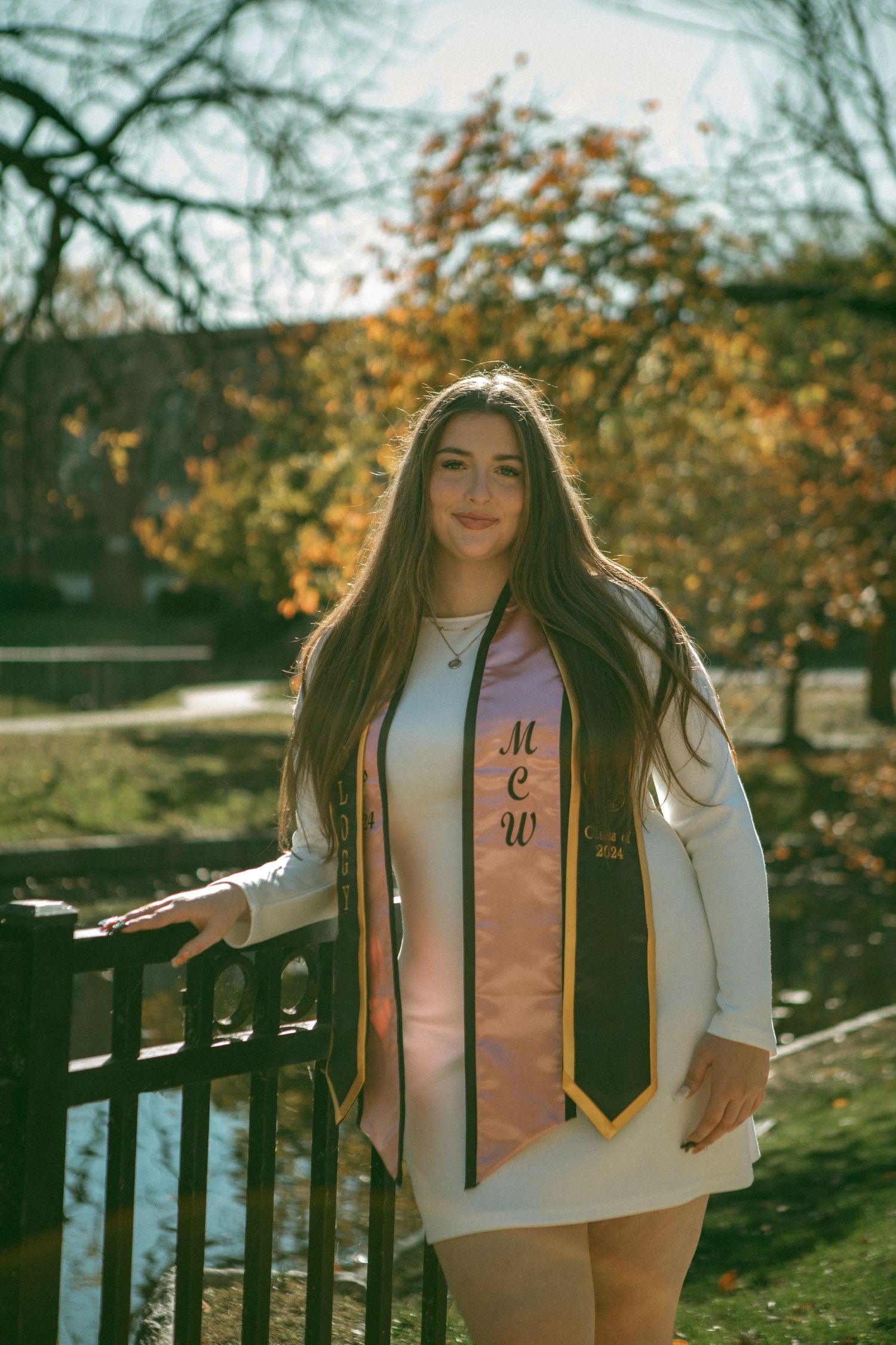 Young woman in white dress wearing two graduation stoles, posing outdoors with autumn leaves in the background, celebrating class of 2024.