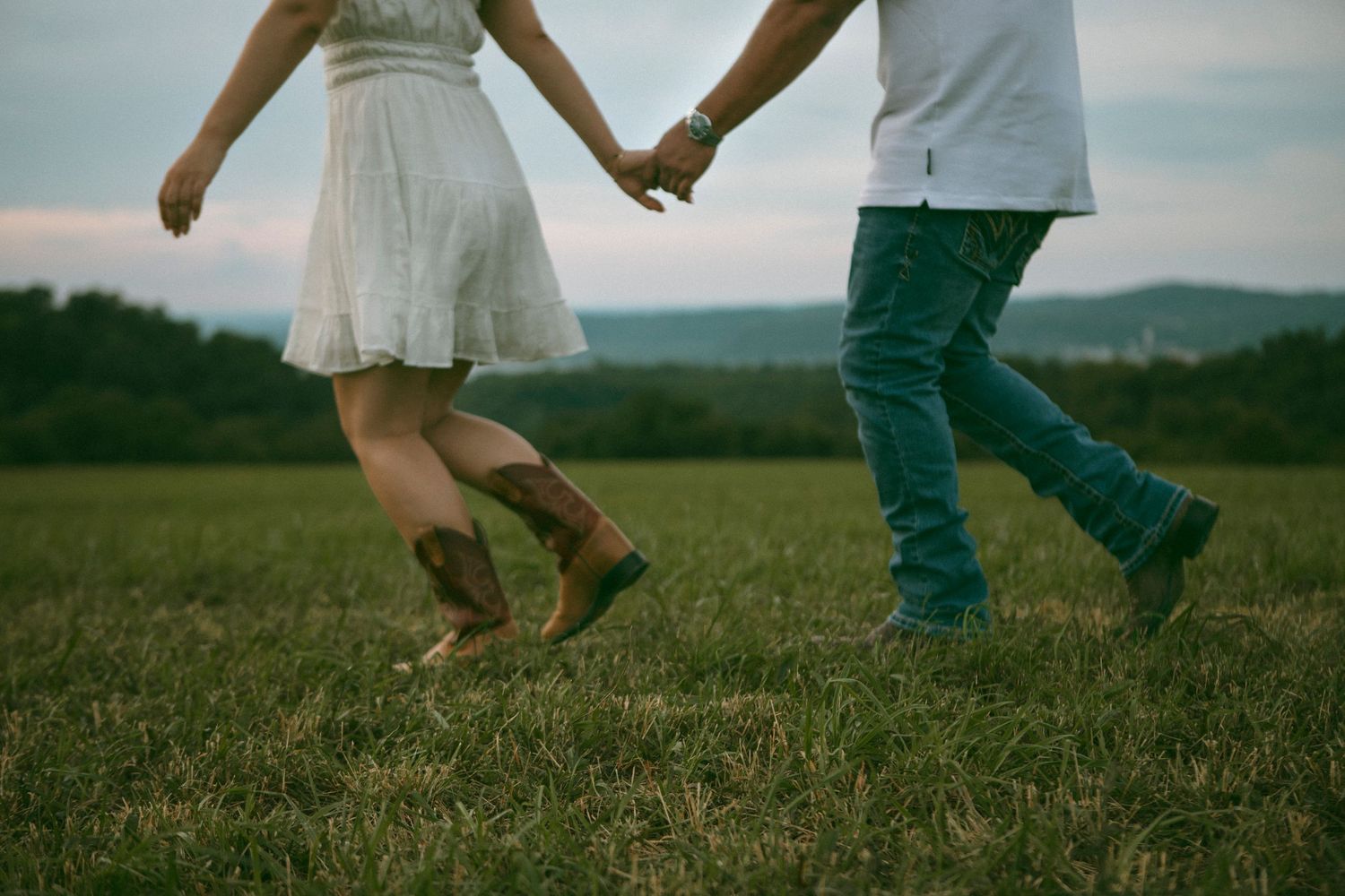 A couple holds hands while jumping together in a grassy field with rolling hills in the background at sunset.