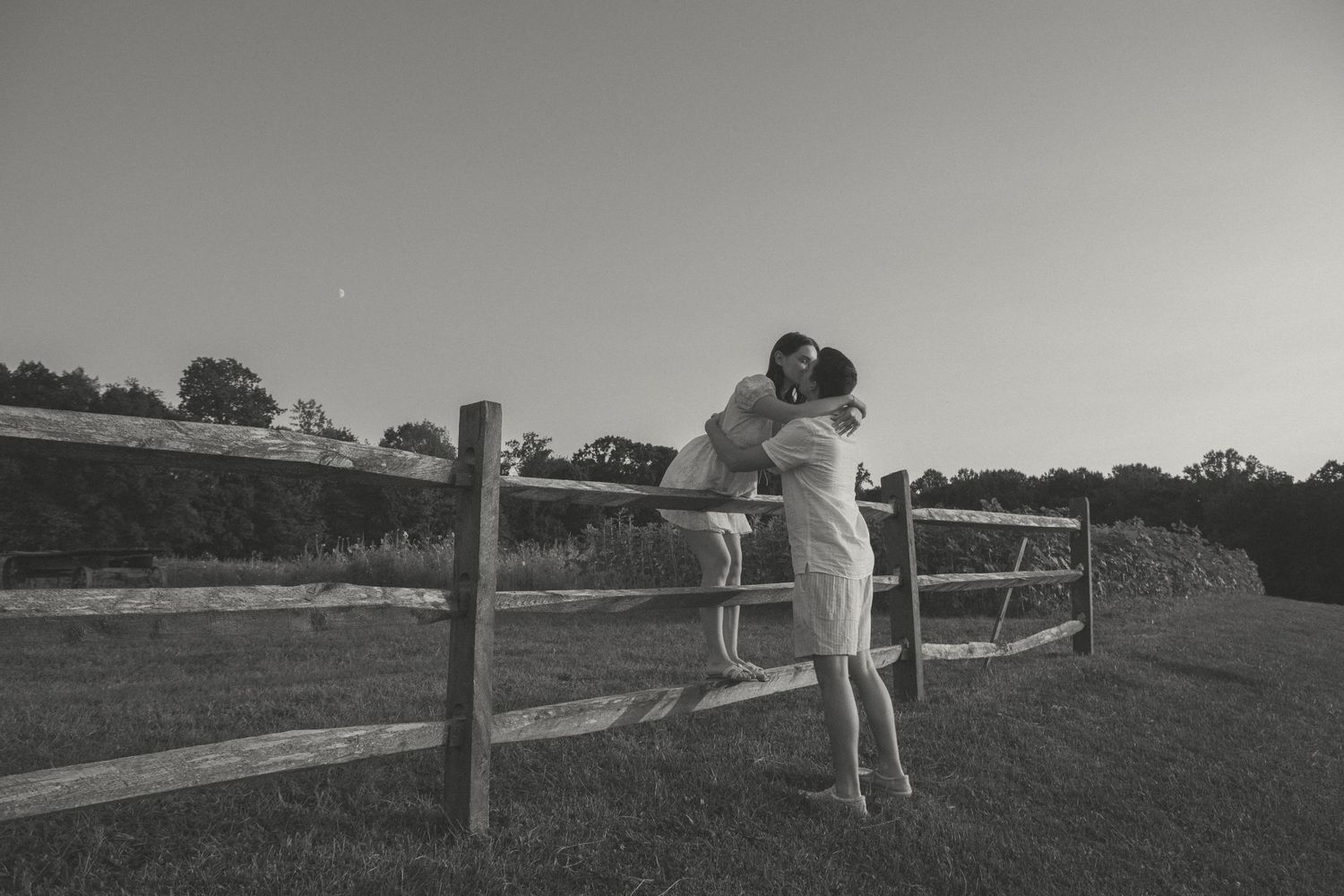 Couple shares a romantic kiss by a rustic wooden fence at sunset, capturing a timeless love story in the countryside.