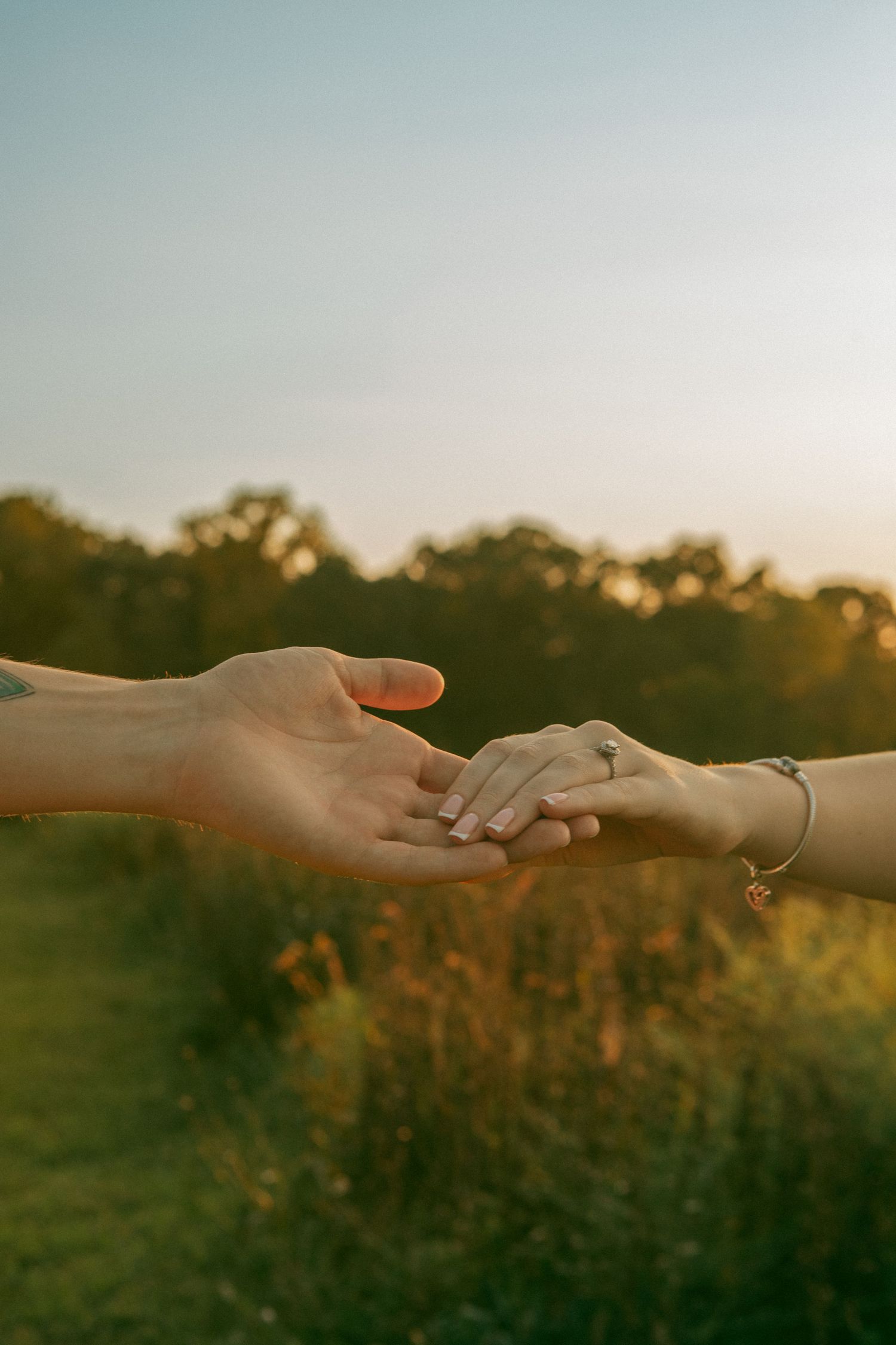 A romantic close-up of a couple’s hands holding each other at sunset, with an engagement ring visible on the woman's hand.