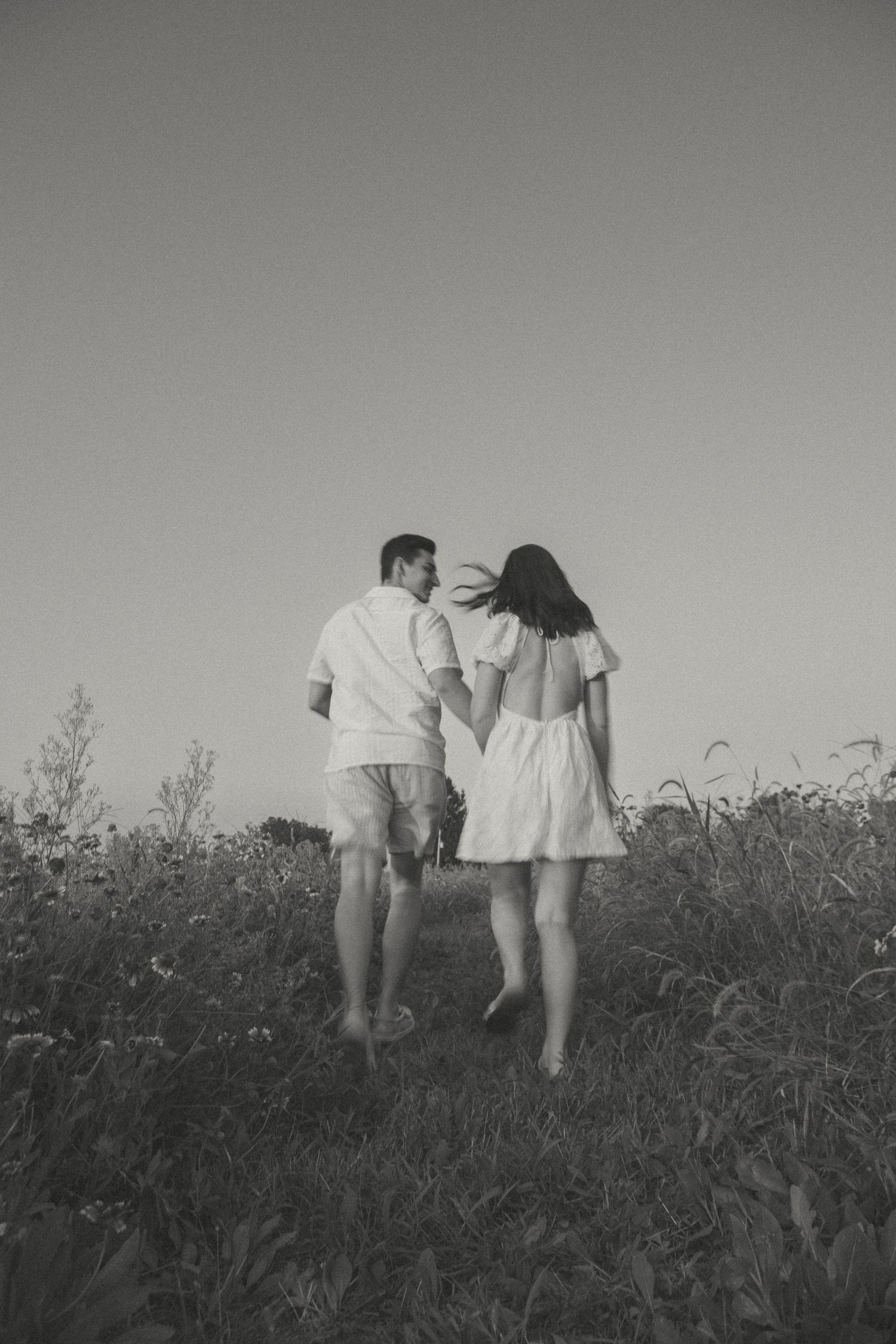 A couple in white outfits walks hand in hand through a wildflower field at sunset, capturing a romantic and timeless moment.
