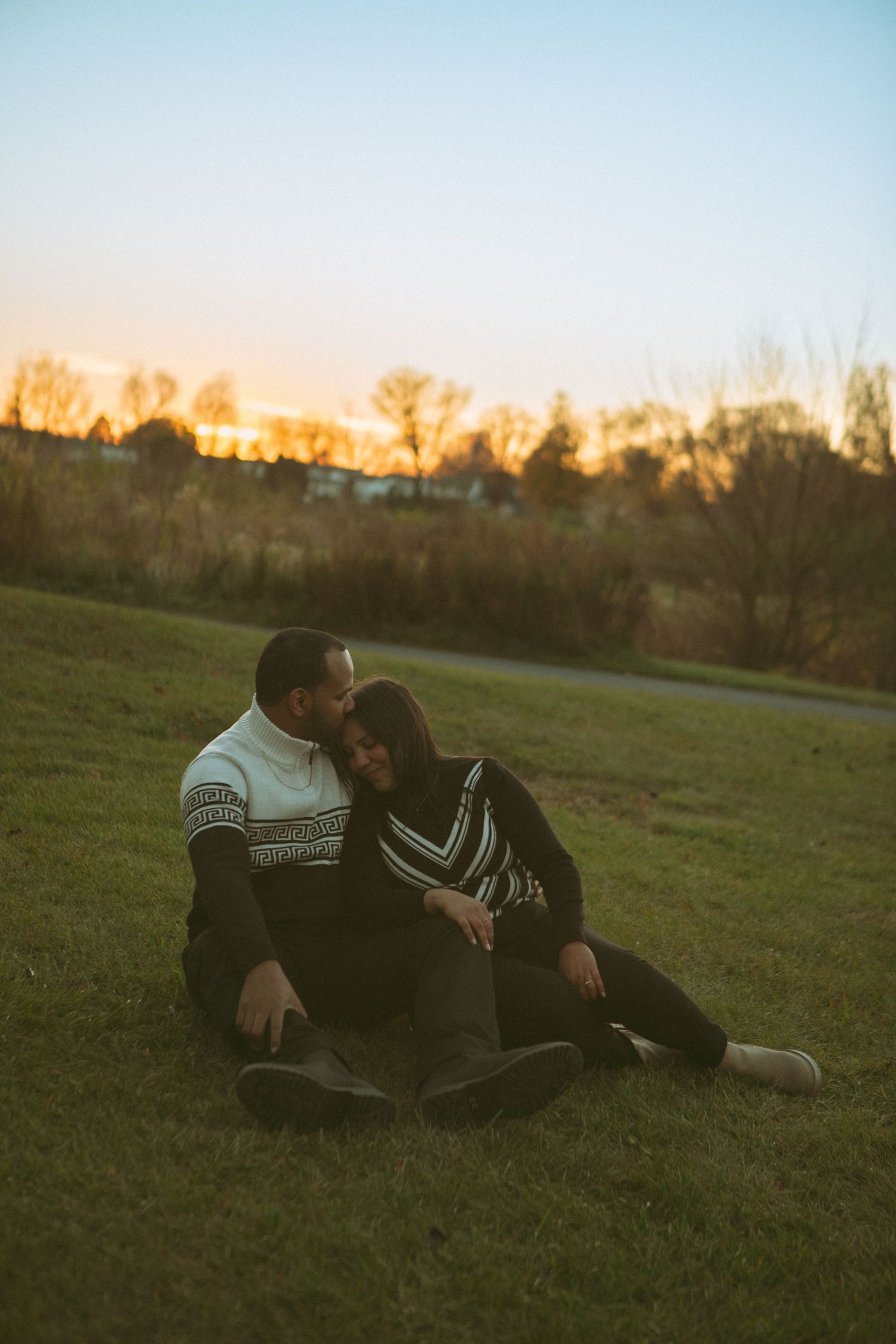 A couple sits on a grassy field at sunset, during a romantic couples photoshoot at Overlook Park in Lancaster, PA