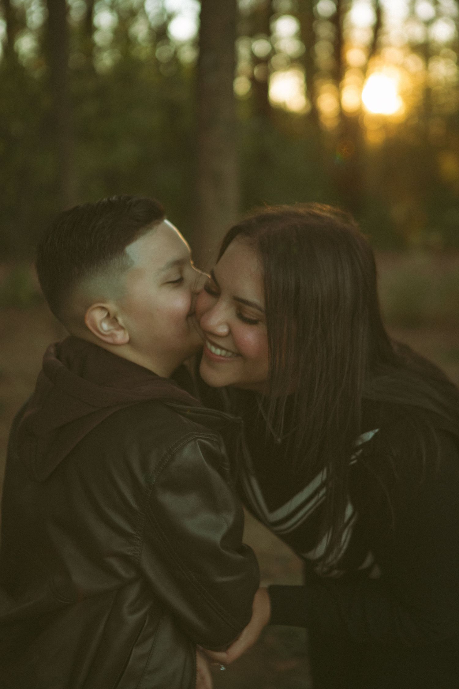 A young boy kisses his smiling mother on the cheek in a golden sunset-lit forest, capturing a heartwarming moment of love and connection.