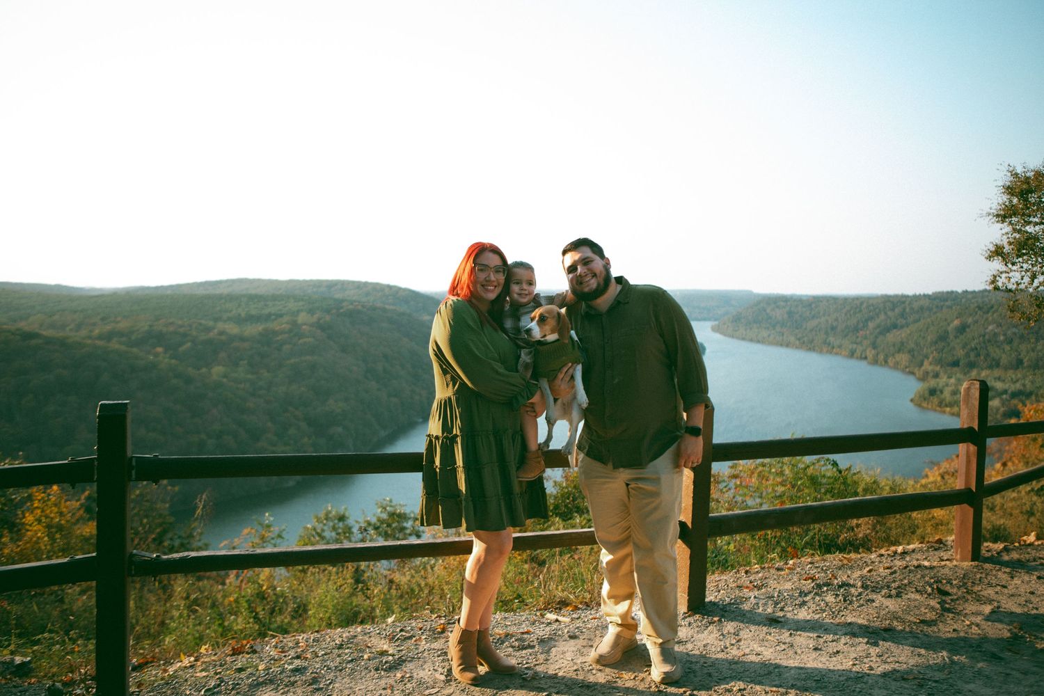 A joyful family and their dog stand together at a scenic overlook, with rolling green hills and a winding river stretching behind them.