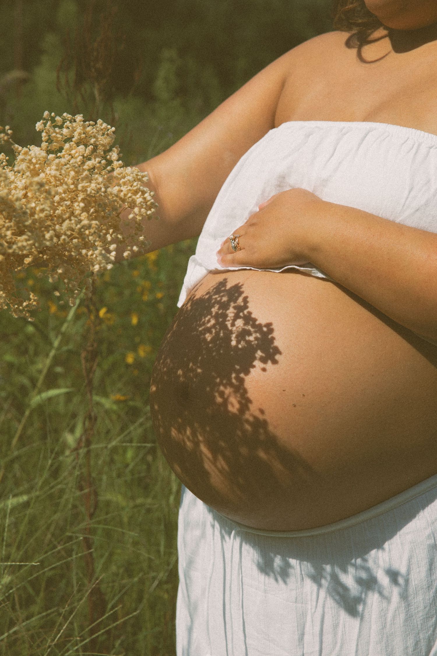 Close-up of a pregnant woman in nature, cradling her belly with floral shadows cast on her skin during maternity session in Lancaster County