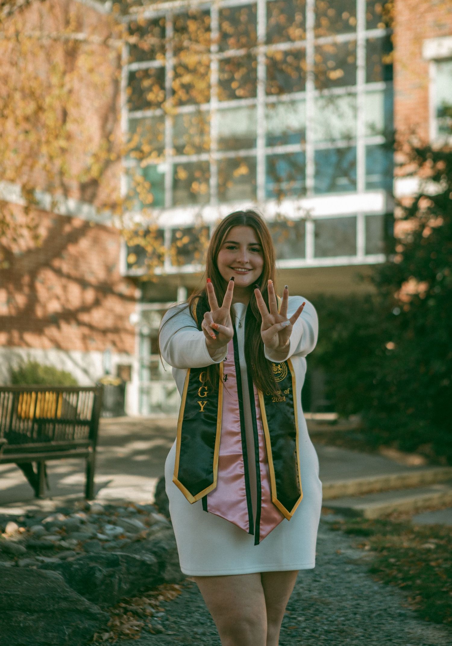 Smiling graduate in a white dress and stole posing with a '24' hand gesture in front of a modern brick building with autumn trees.