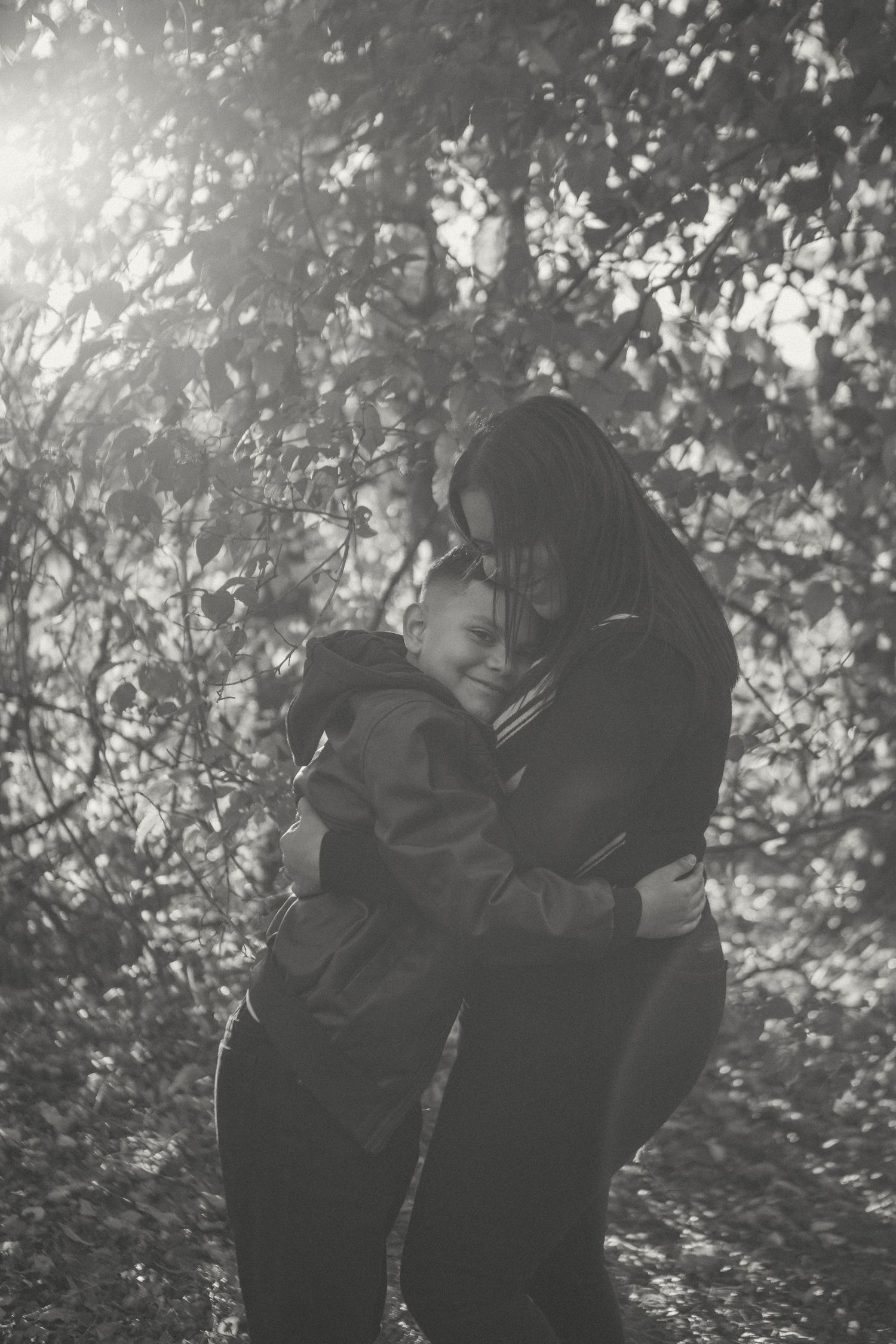 A mother and son share a warm embrace in a sunlit forest, captured in a black and white portrait filled with love and connection.