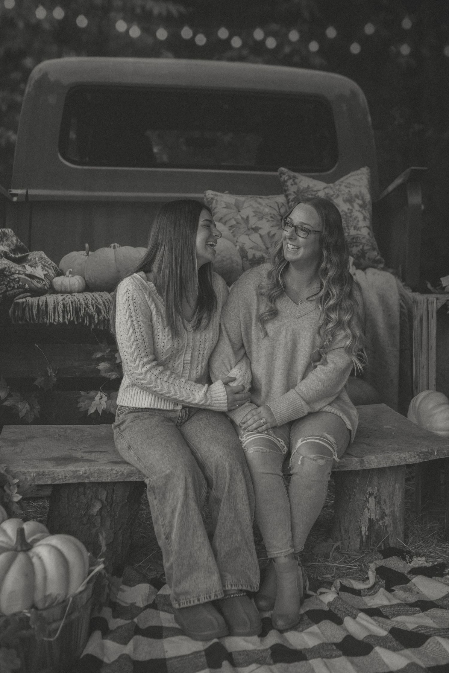 Mother and daughter sit on a rustic bench, laughing and holding hands, with pumpkins and a vintage truck in the autumn-themed background.