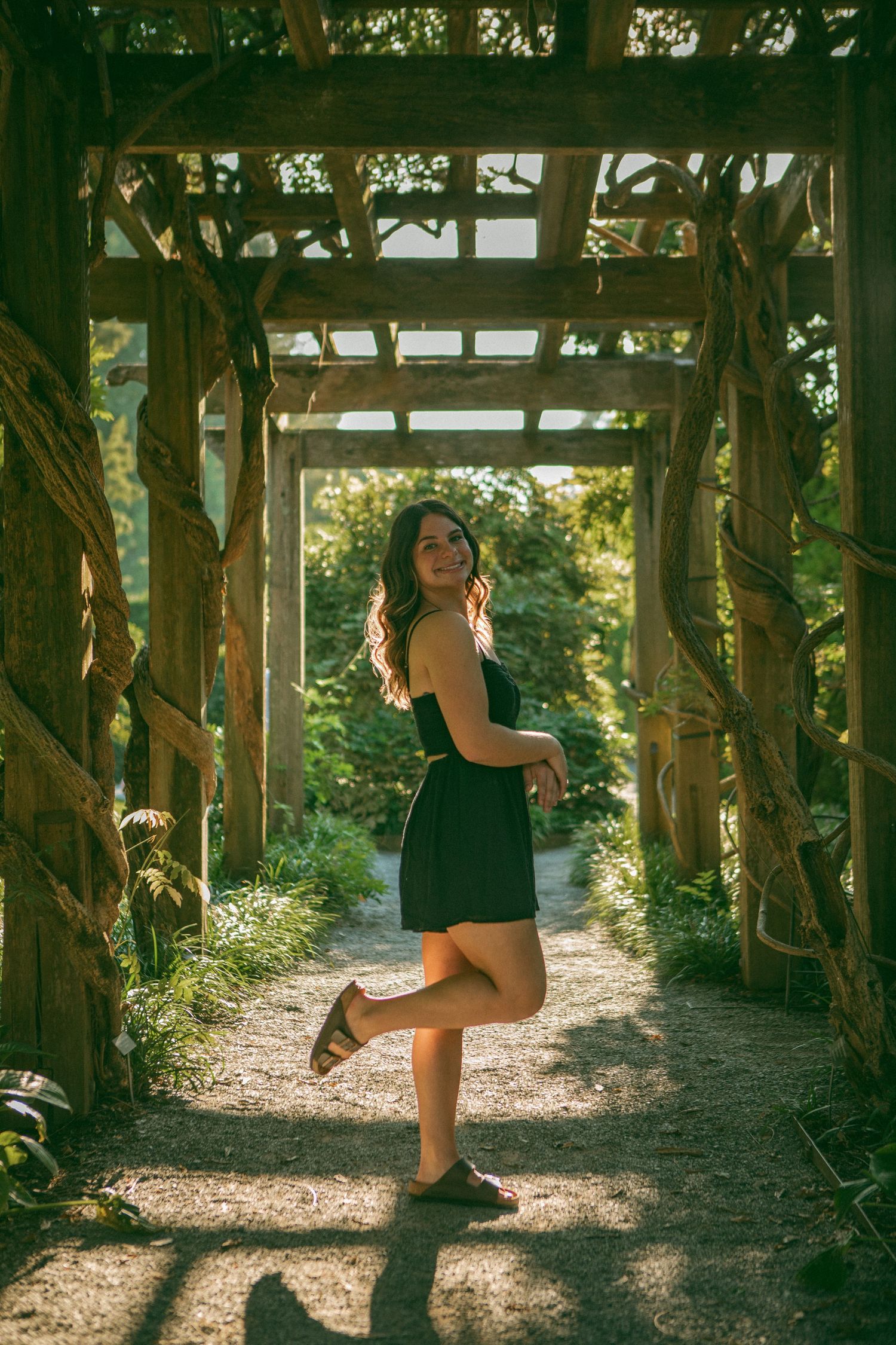 Young woman in a black dress poses playfully under a rustic wooden pergola entwined with vines, bathed in warm golden sunlight.