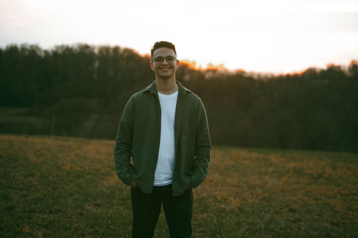 A male from a couples session at Highpoint Scenic Vista in Wrightsville stands alone in grassy field at sunset with trees in background.