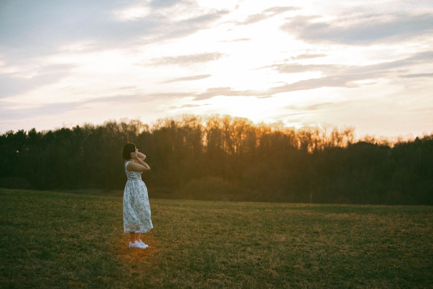 A woman from a couples session in Wrightsville, PA, looks away at sunset while golden light streams through bare trees on the horizon.