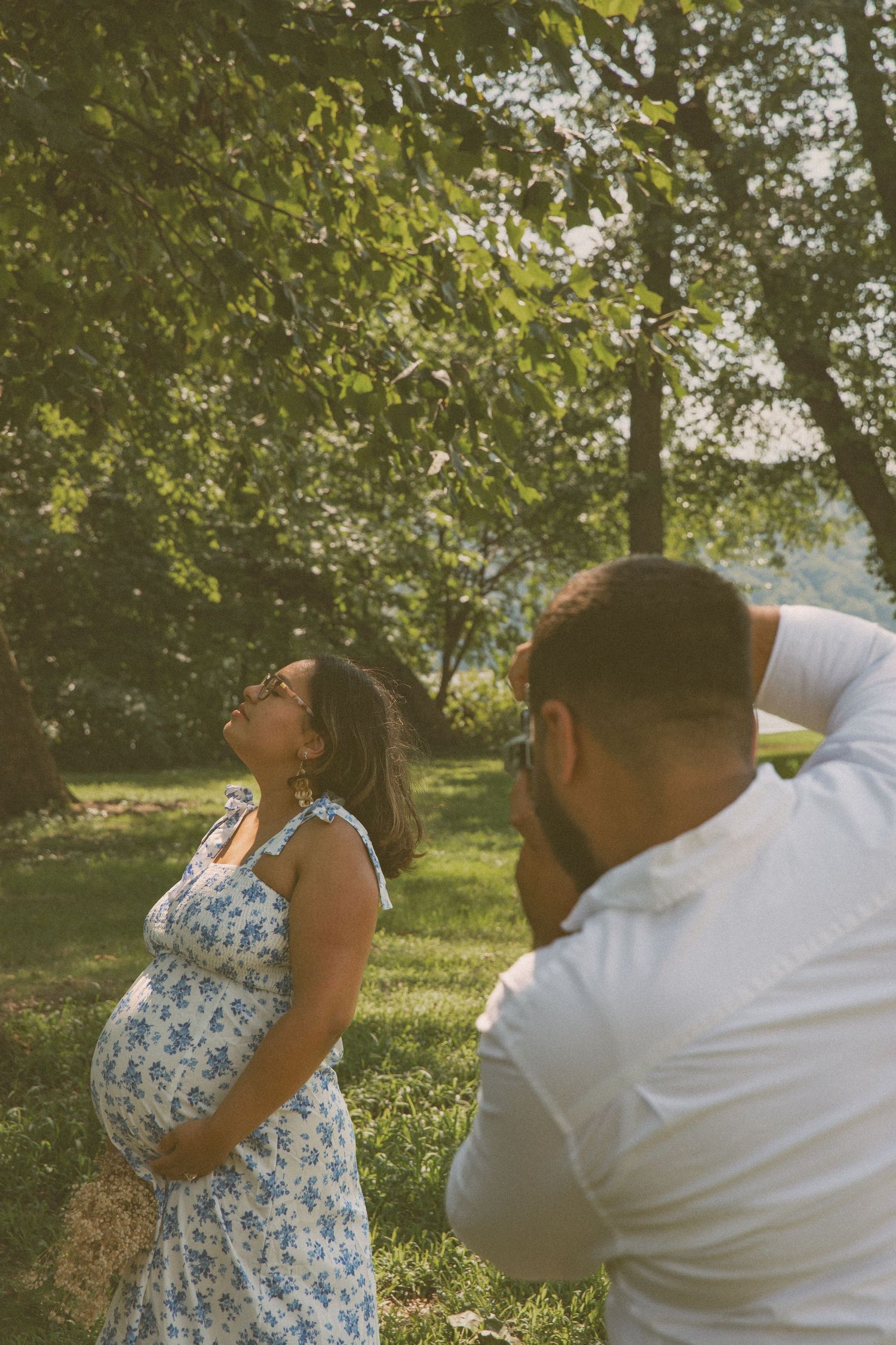 Pregnant woman poses in a floral dress as her partner captures the moment with a camera in a lush, green outdoor setting with natural light.