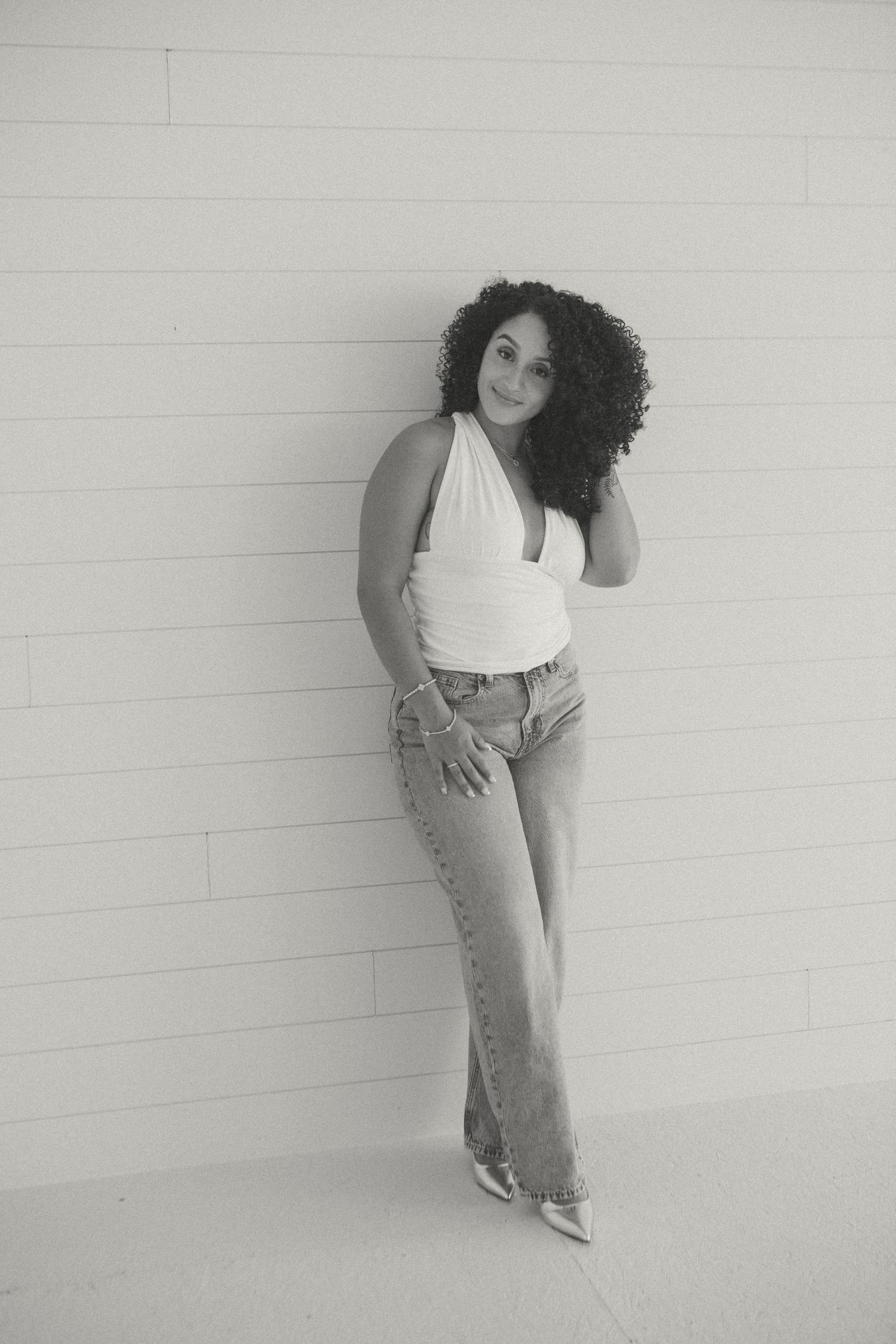 A woman with curly hair leans against a white shiplap wall, wearing a halter top and jeans, exuding effortless confidence.
