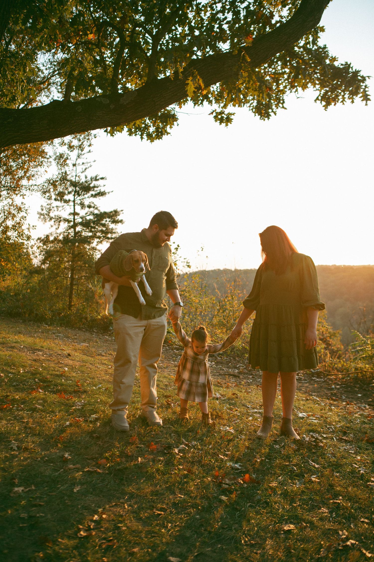 Family walks at sunset, holding hands with their child while the father carries their dog, capturing a warm and loving outdoor moment.