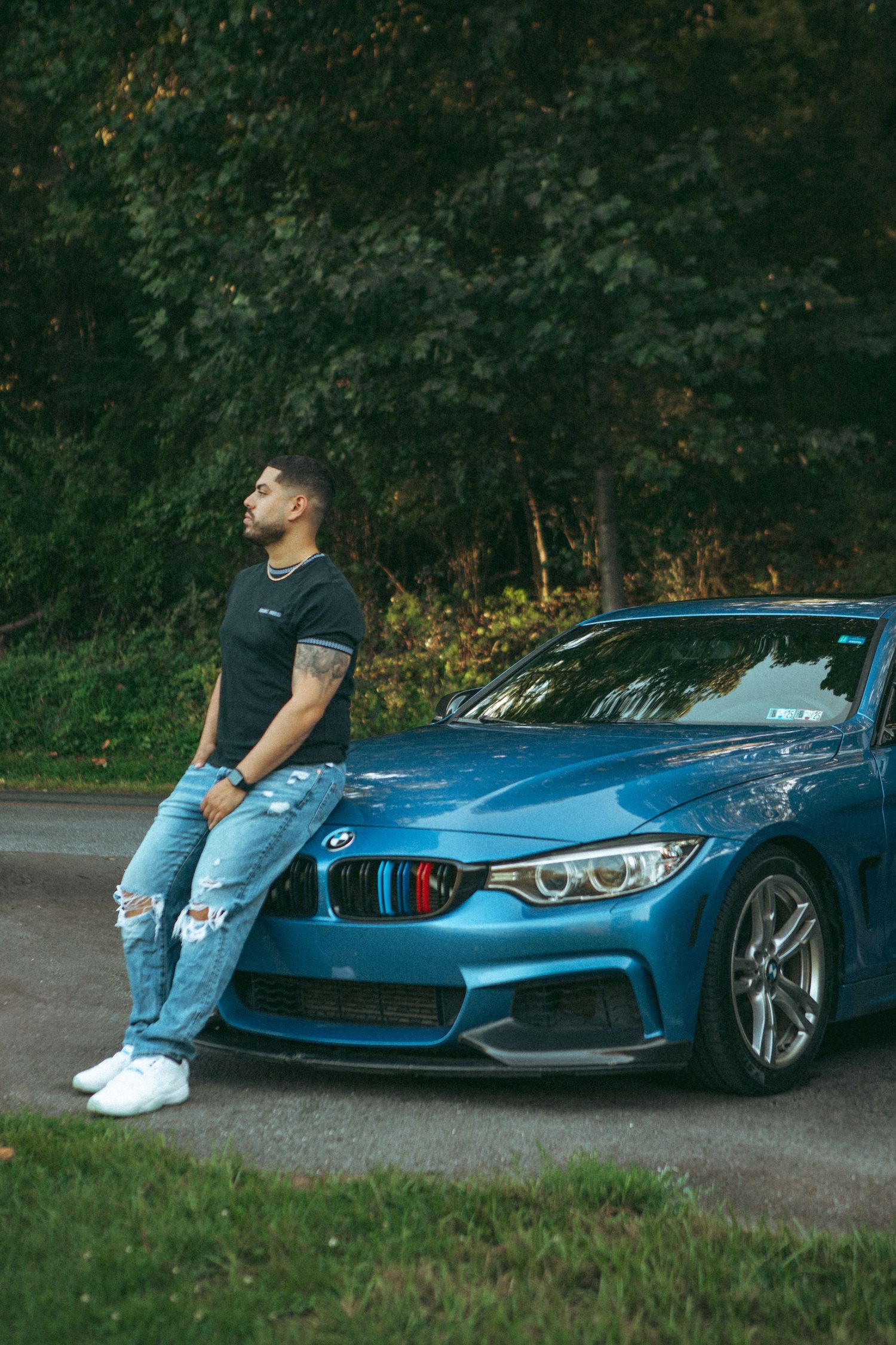 A man leaning against a blue BMW, dressed in a black t-shirt and ripped jeans, with a relaxed expression, set against a backdrop of greenery