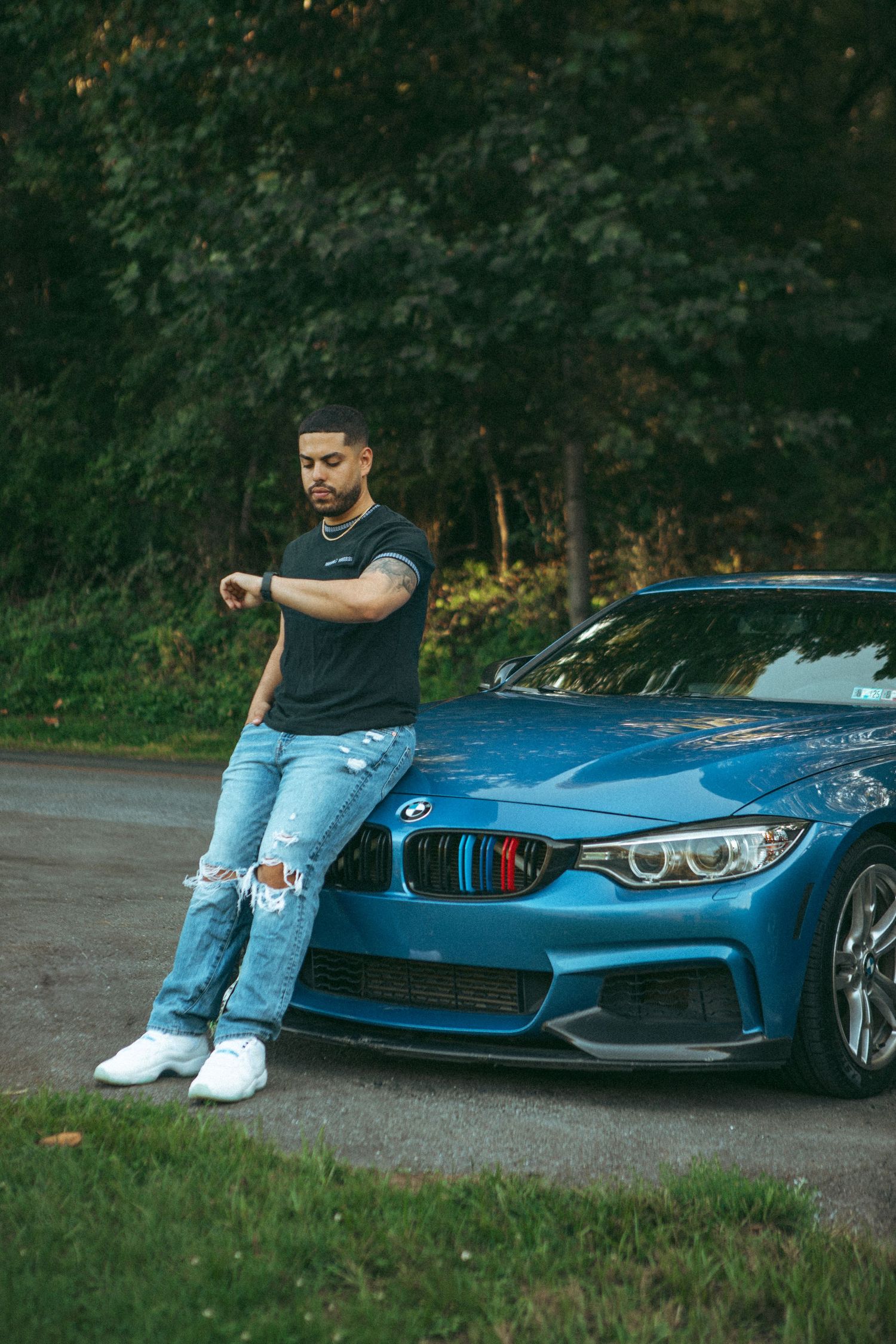 Stylish man sitting on a blue BMW, checking his watch – urban automotive lifestyle photography with a scenic backdrop.