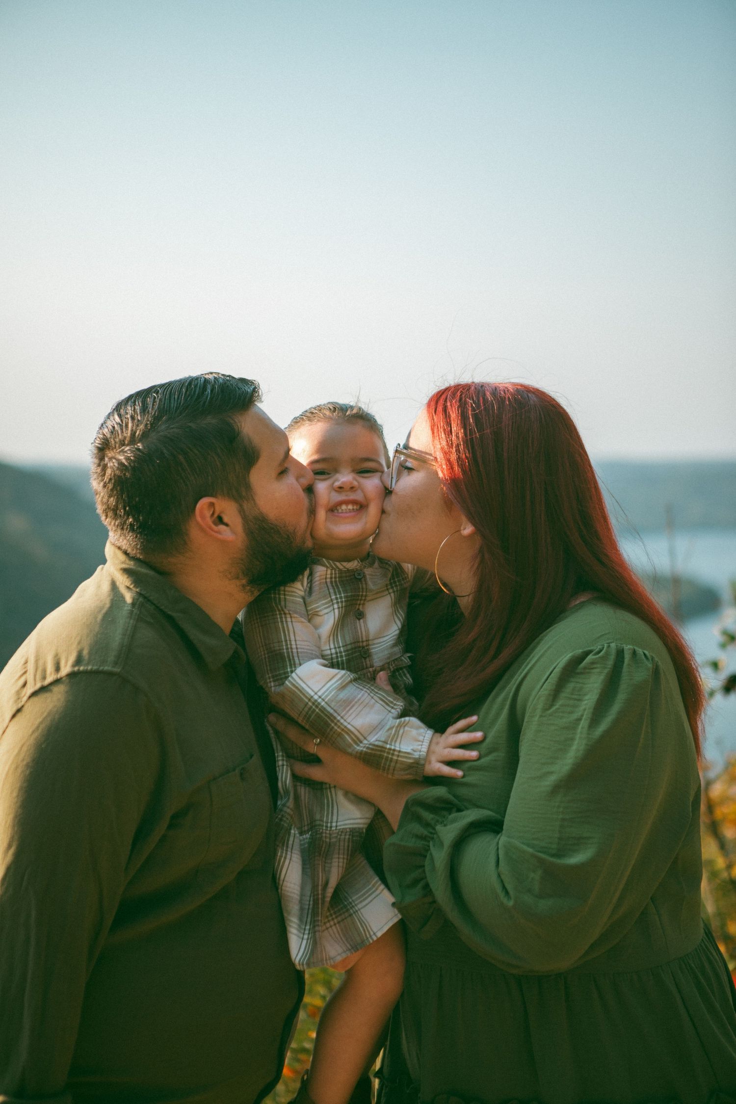 Parents kiss their smiling daughter on the cheeks with a scenic lake view in the background, capturing a heartwarming family moment.