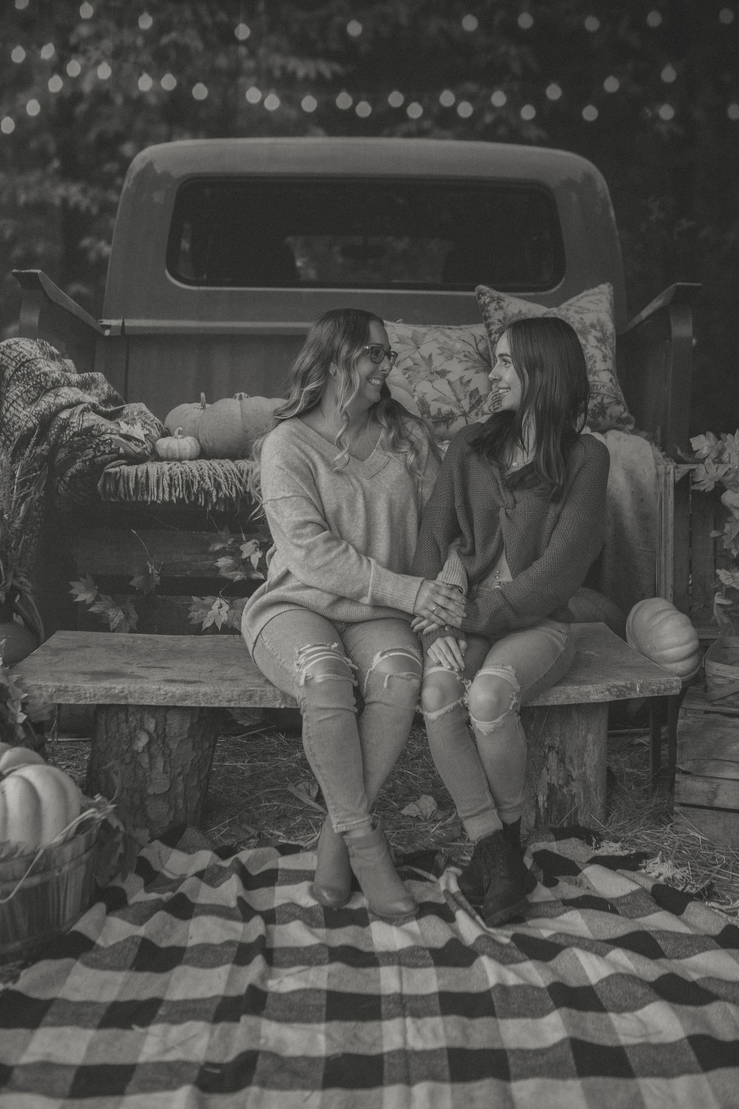 Mother and daughter sit on a rustic bench, holding hands and smiling, with an autumn-themed setup behind them, featuring a vintage truck.