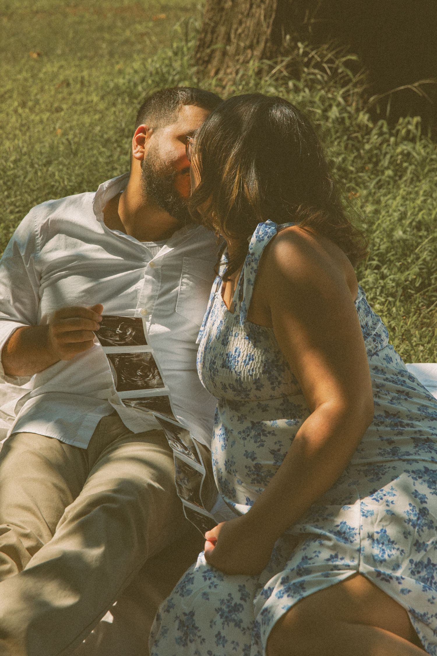 Expecting couple shares a loving moment outdoors, holding ultrasound photos as they celebrate their pregnancy in a dreamy, sunlit setting.