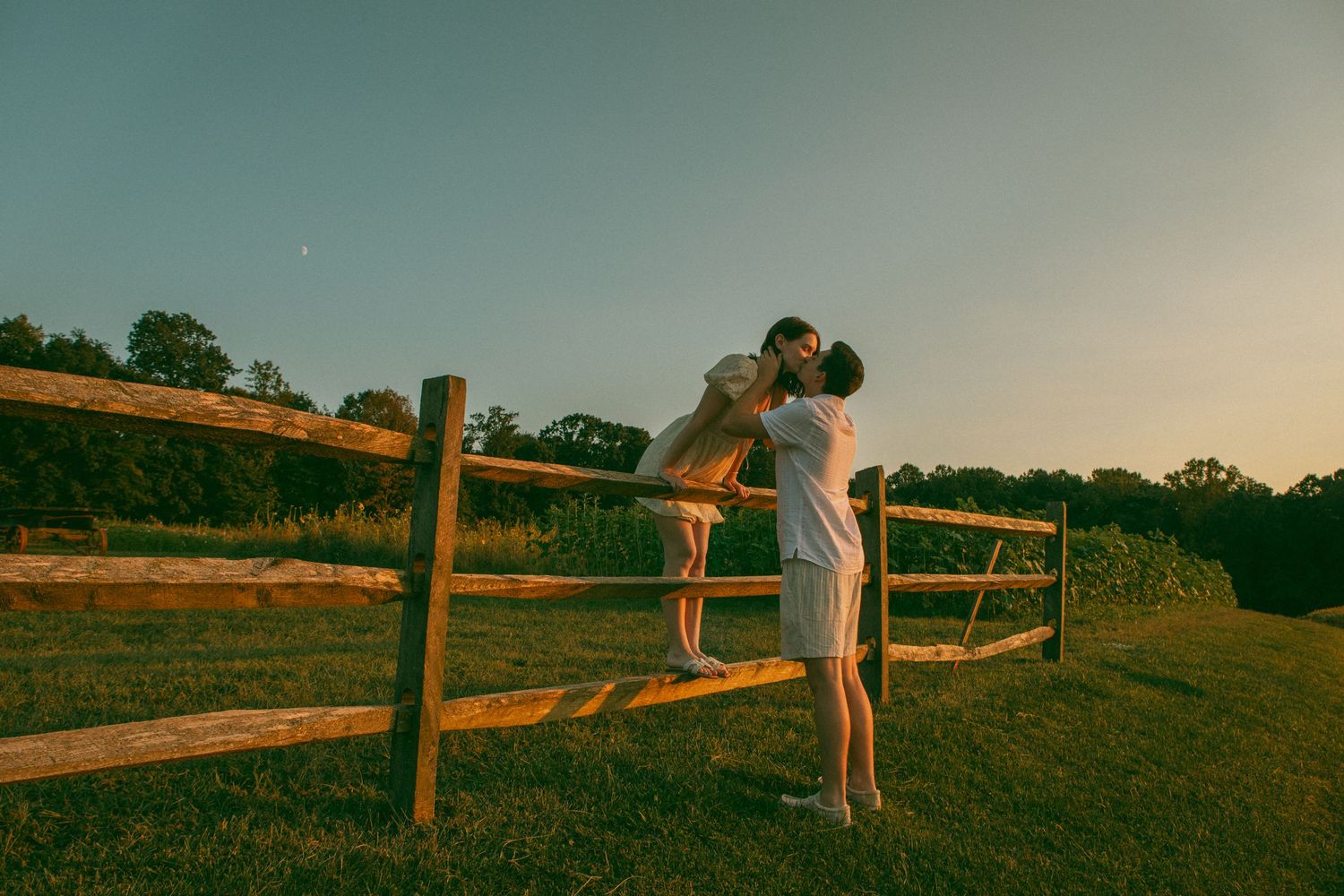 Couple sharing a romantic kiss over a rustic wooden fence at sunset, bathed in golden light, evoking warmth, love, and countryside charm.