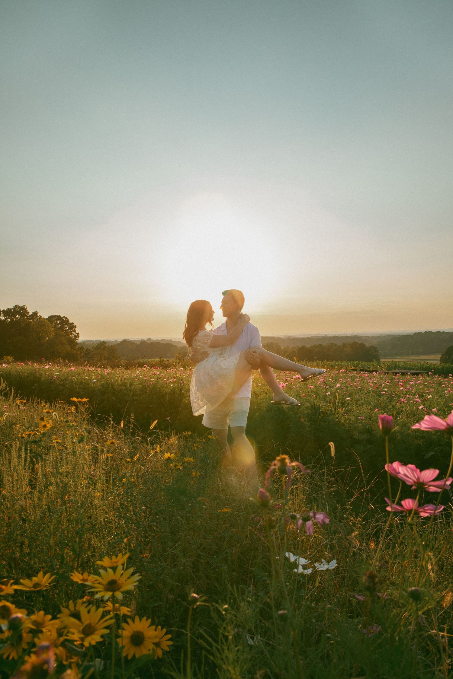 Man lifting his partner in a blooming wildflower field at sunset, creating a romantic, golden-hour moment filled with love and warmth.